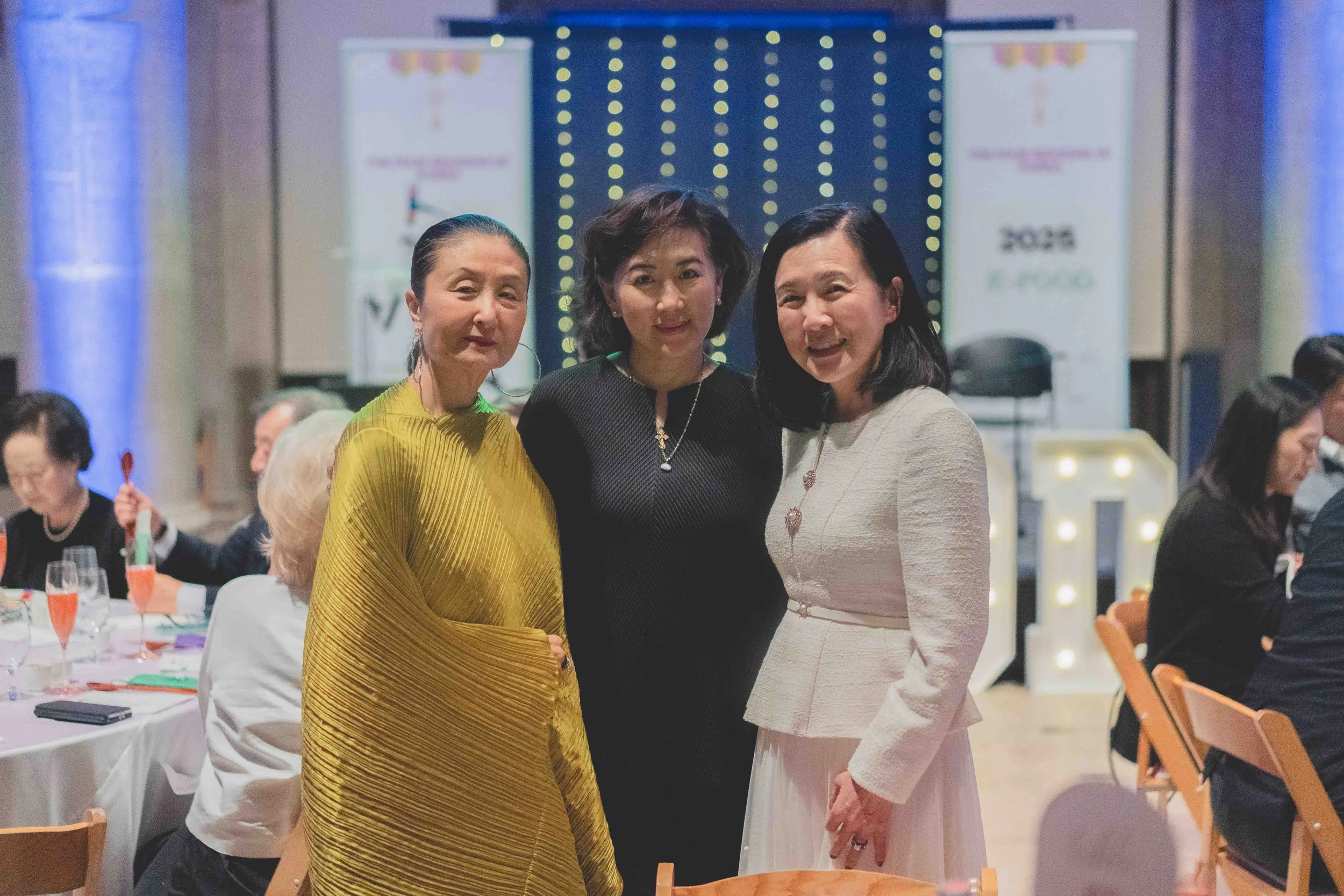 Three women standing together at a formal event, all smiling and dressed in elegant attire, with other guests seated at tables in the background.