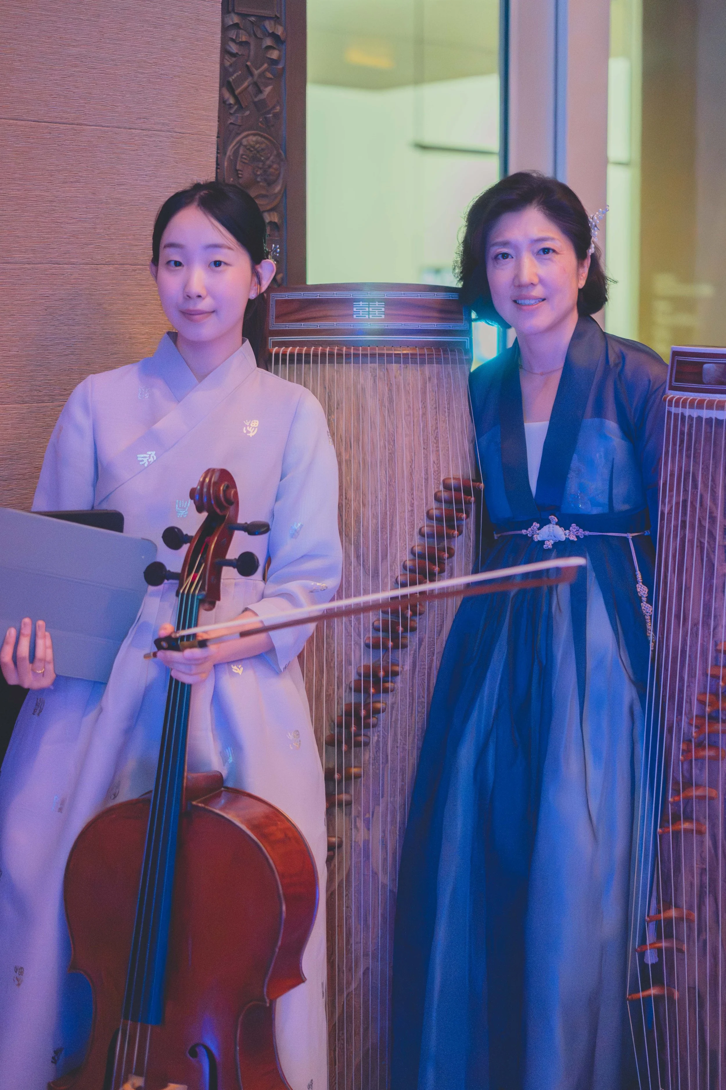 Two women, one holding a violin and bow, standing in front of a traditional wooden Korean harp, with decorative woodwork and a bright screen in the background.