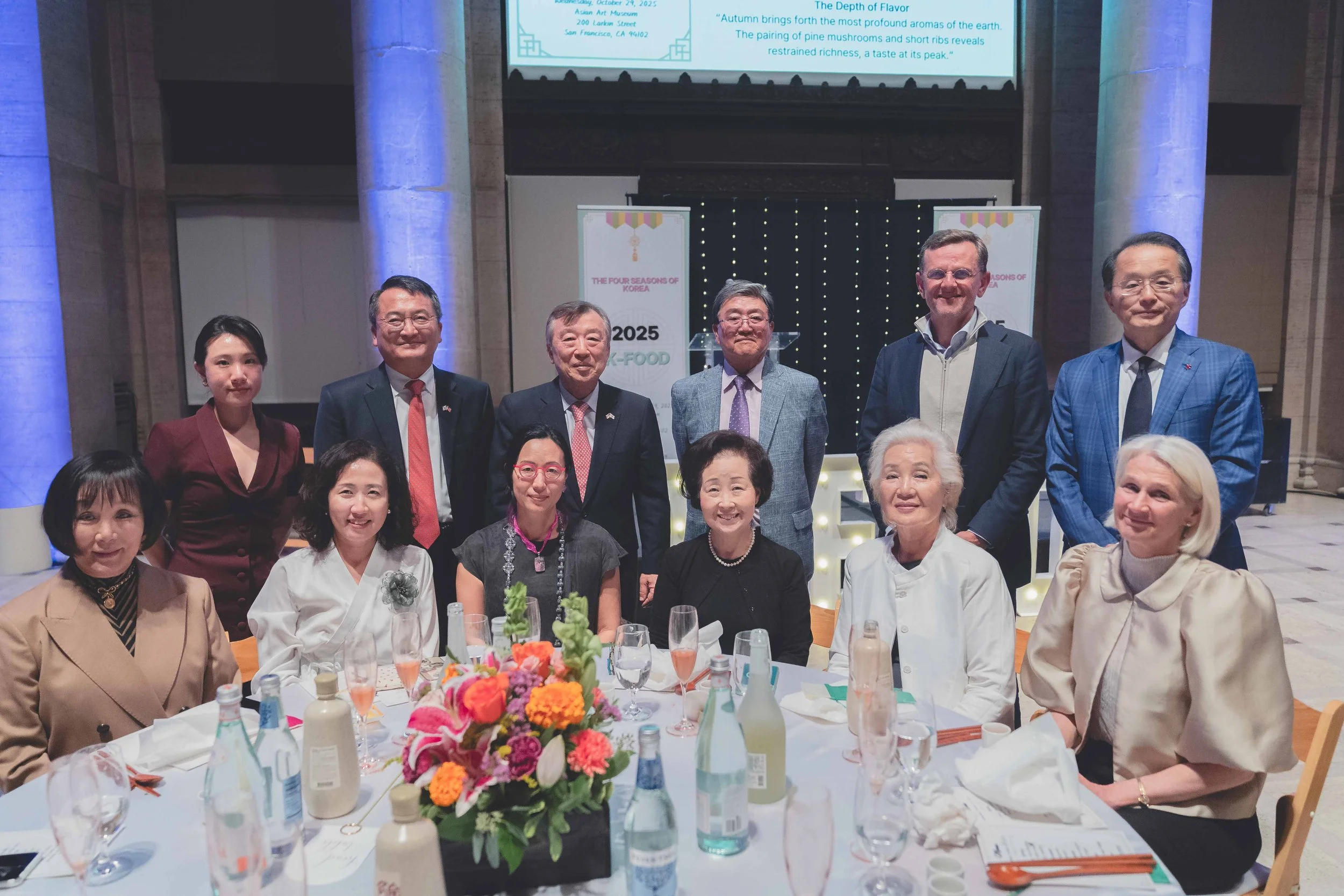 Group of people at a formal event, seated and standing around a round table with floral centerpiece and drinks, in a decorated indoor venue with banners and a large screen in the background.