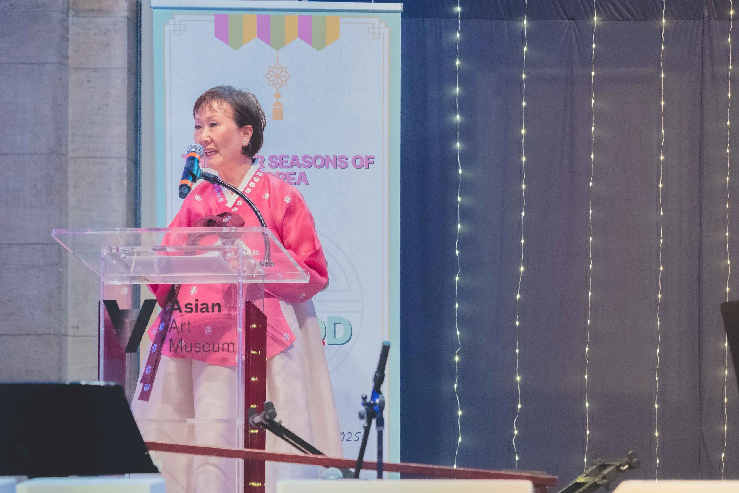 A woman in traditional Korean attire speaking at a podium at the Asian Art Museum event, with a colorful backdrop and fairy lights on the right side.