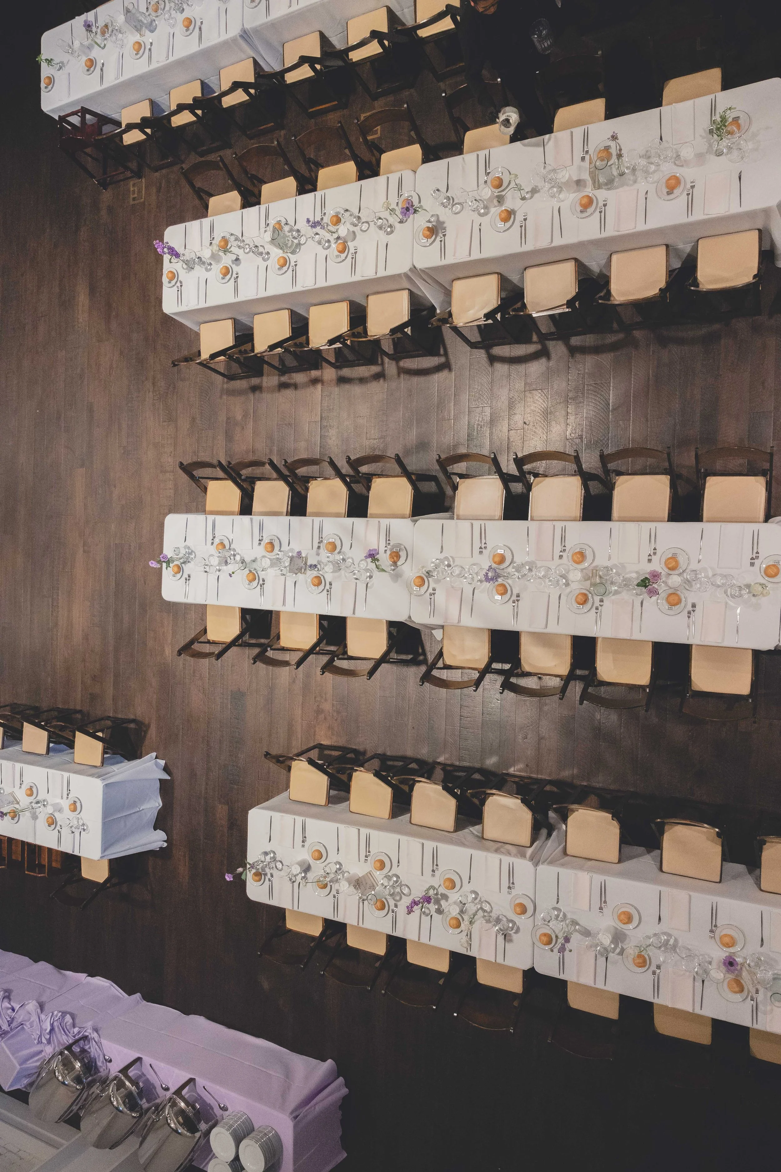 Aerial view of a banquet hall setup with long tables covered in white tablecloths, neatly arranged chairs, and table settings including plates, cutlery, and glassware. Tables are adorned with floral centerpieces.