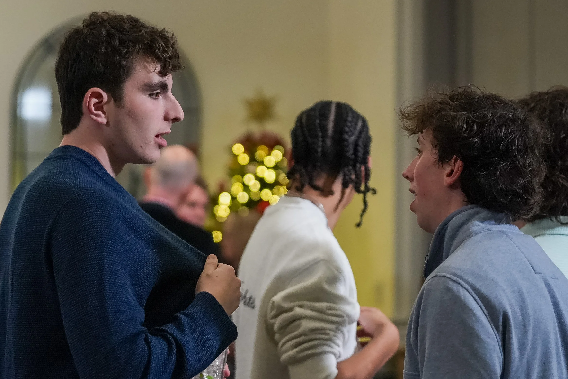 Group of people socializing indoors with a blurred Christmas tree in the background.