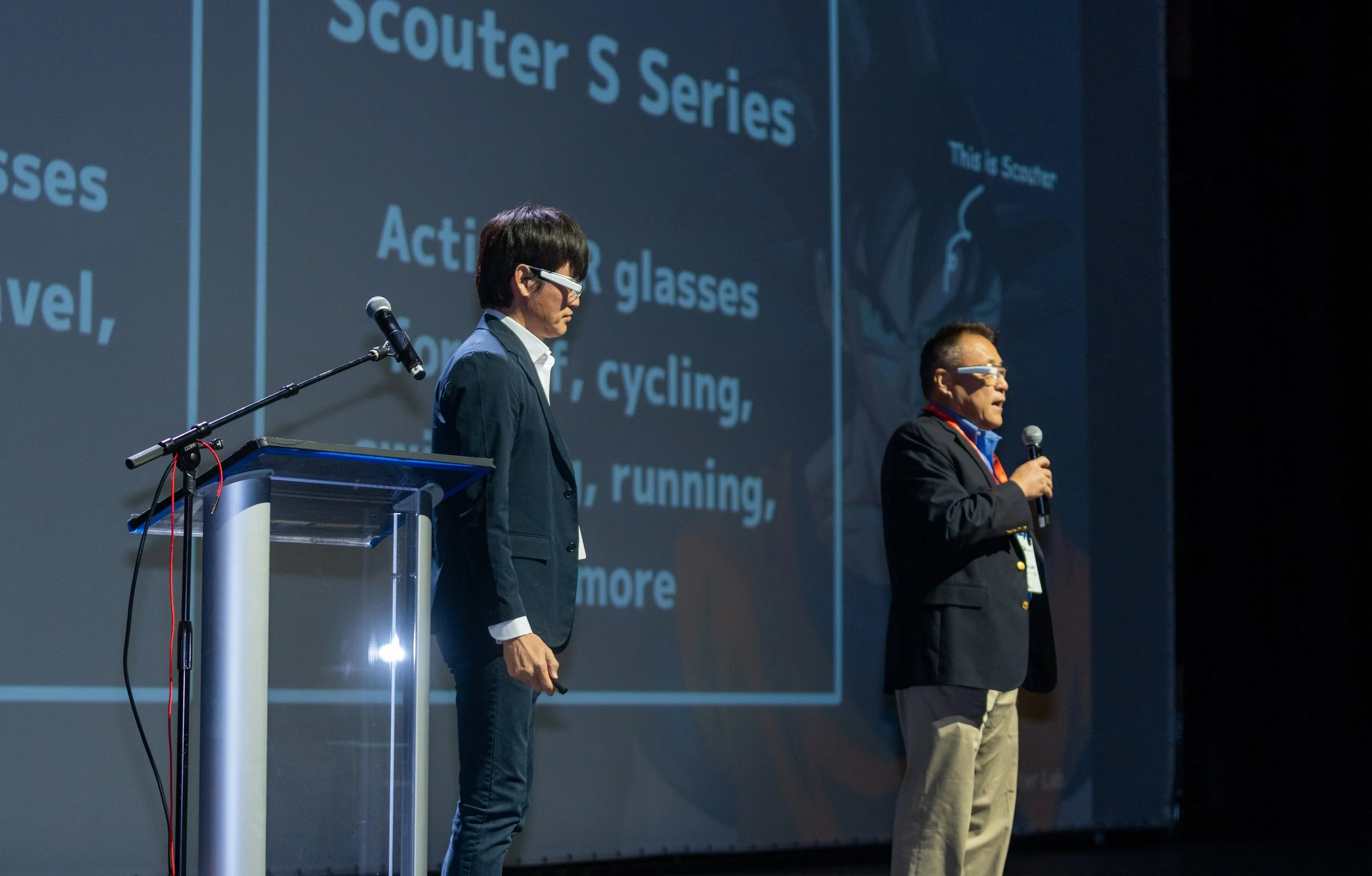 Two men on stage presenting with microphones, wearing smart glasses, with a large screen behind them displaying text about the "Scouter S Series" glasses for various activities.