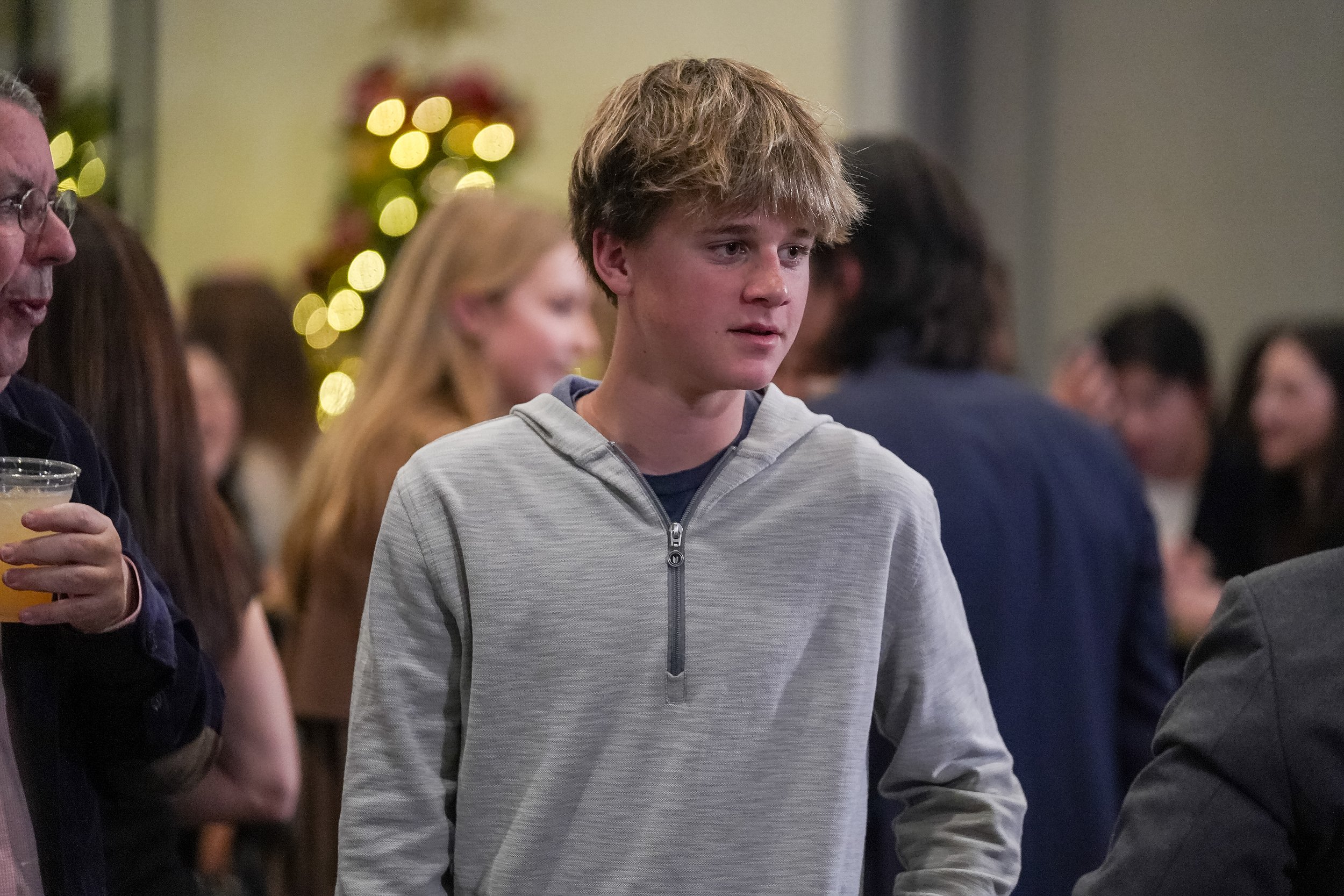 Young person in gray hoodie at a social gathering with blurred people in the background.