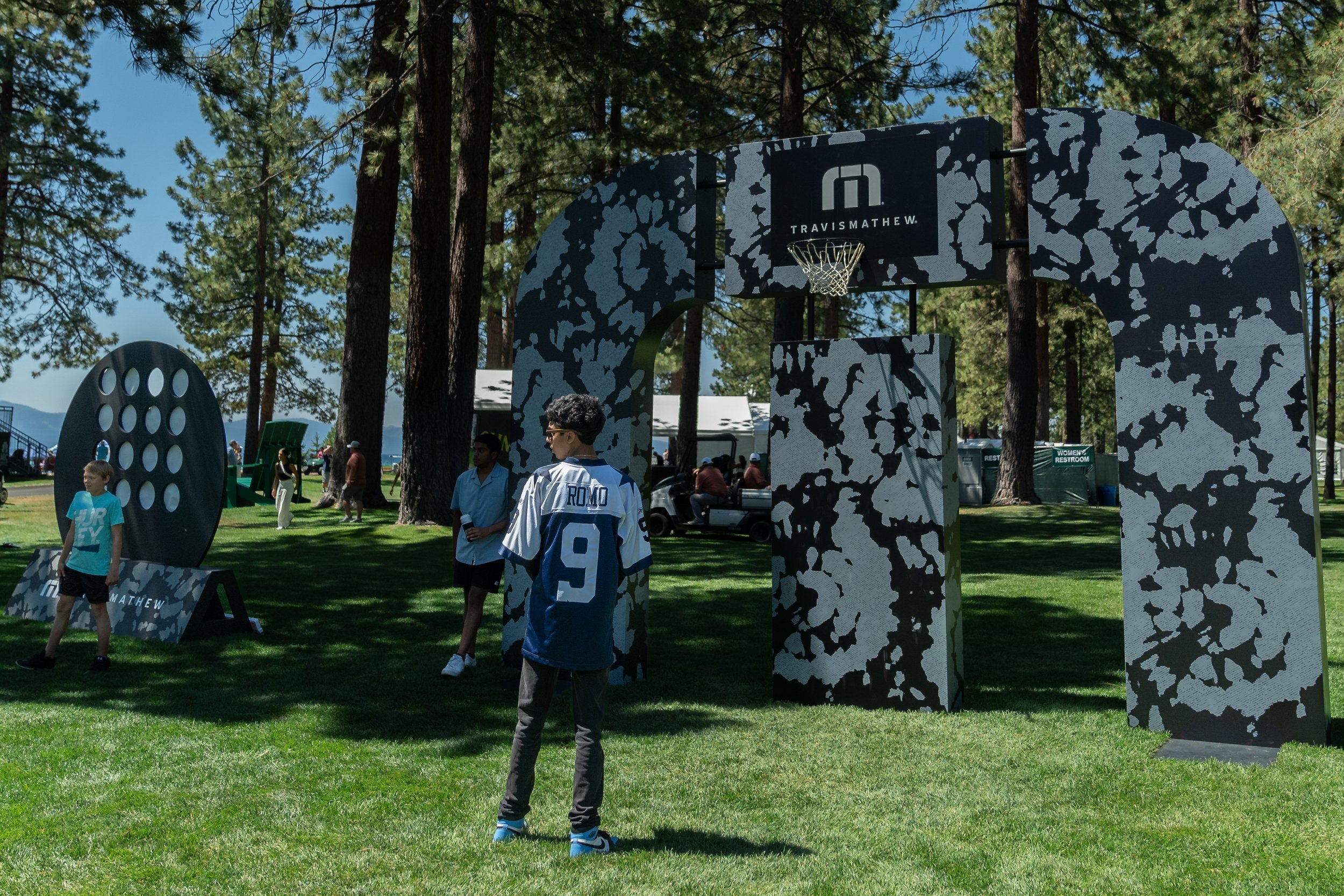 Outdoor event with camouflage-patterned arches and Travis Mathew branding, people in casual attire playing games on a grassy area, surrounded by tall trees.