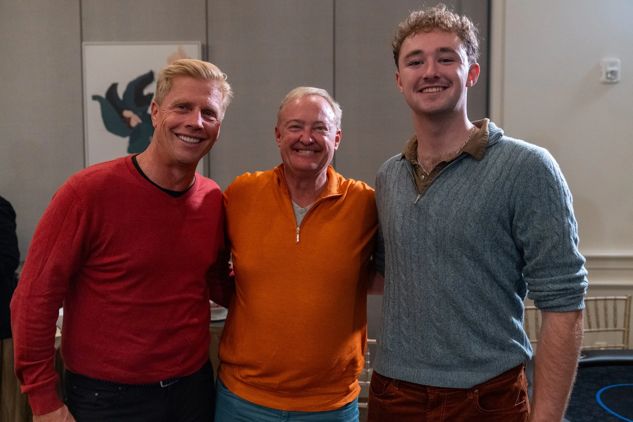 Three smiling men standing together indoors, one in a red sweater, another in an orange sweater, and the third in a blue sweater. They are in front of a wall with a framed artwork.