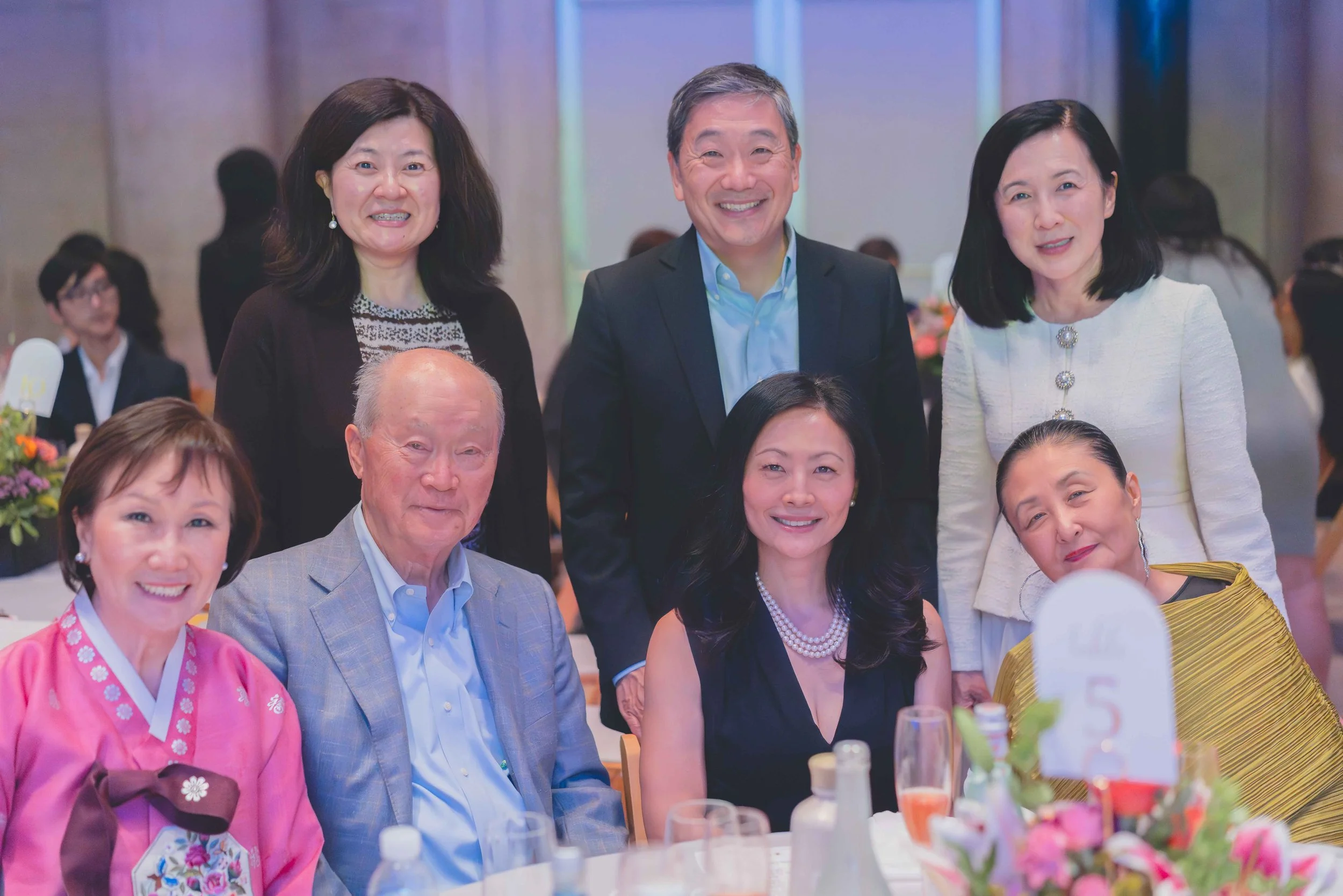 A group of seven people, five women and two men, gathered at a formal event, smiling at the camera. They are dressed in colorful and business attire, sitting and standing around a table with flower arrangements and beverages in a banquet hall.