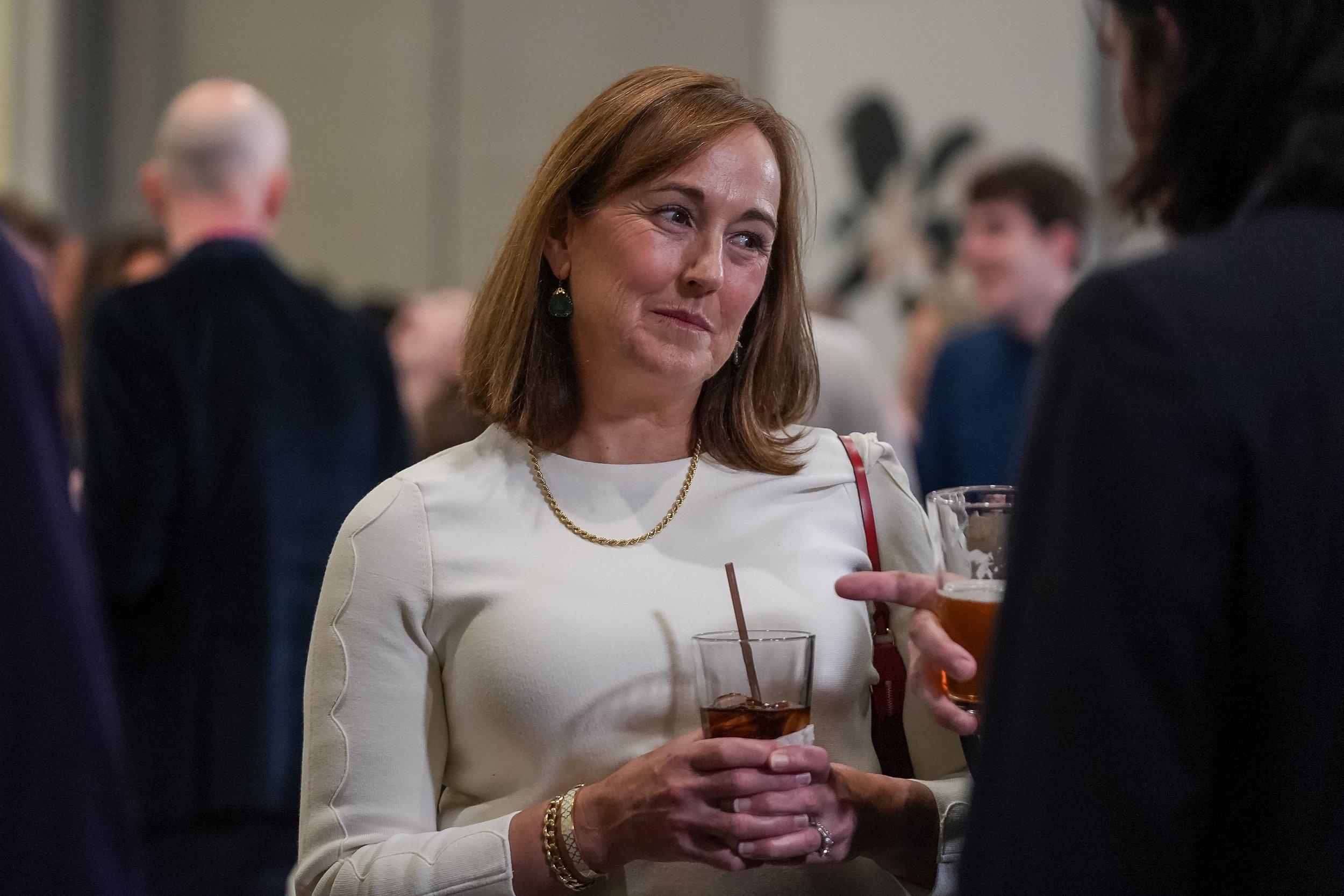 Woman in white dress holding a drink, engaged in conversation at a social event.