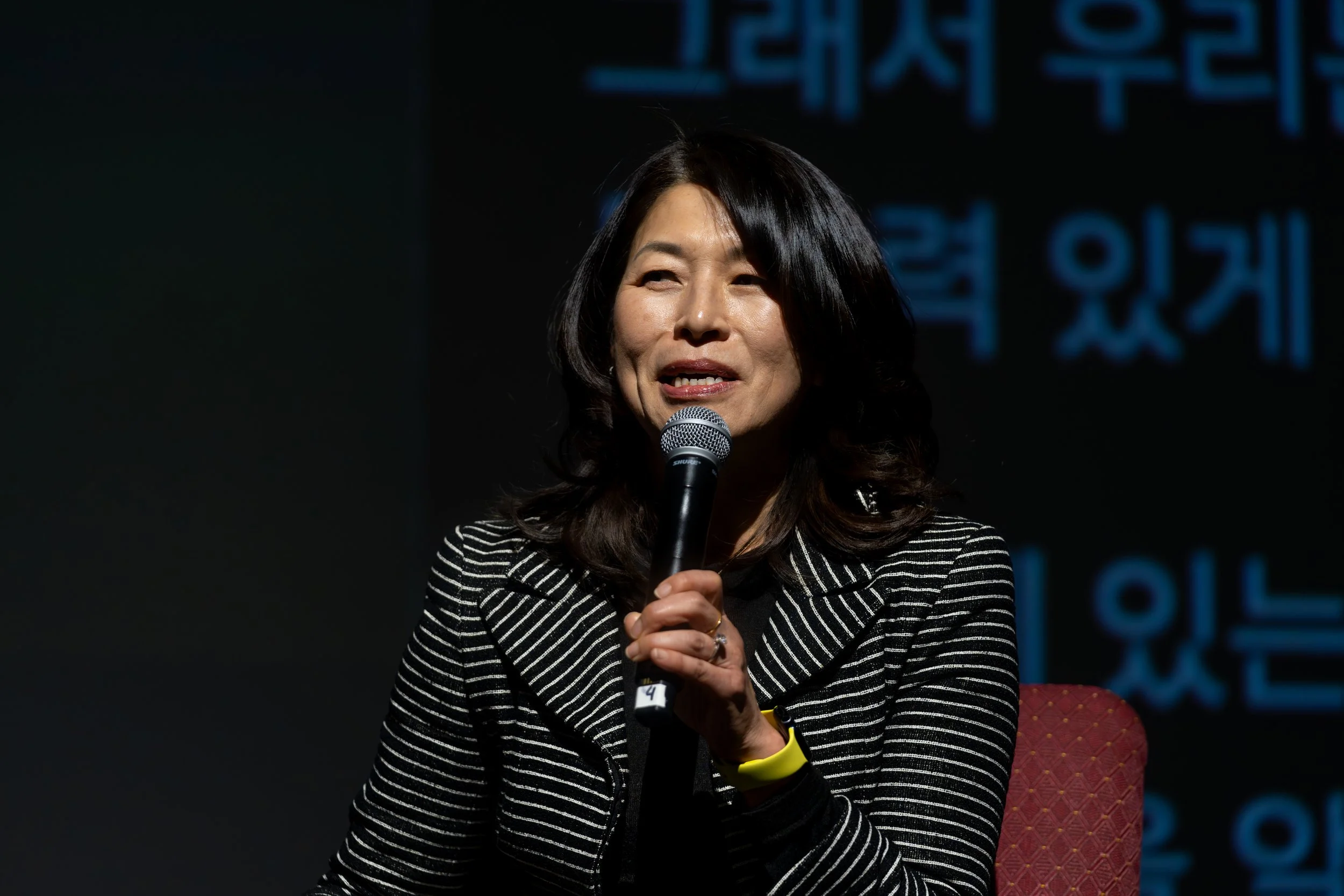 Woman speaking into a microphone, wearing a striped blazer.