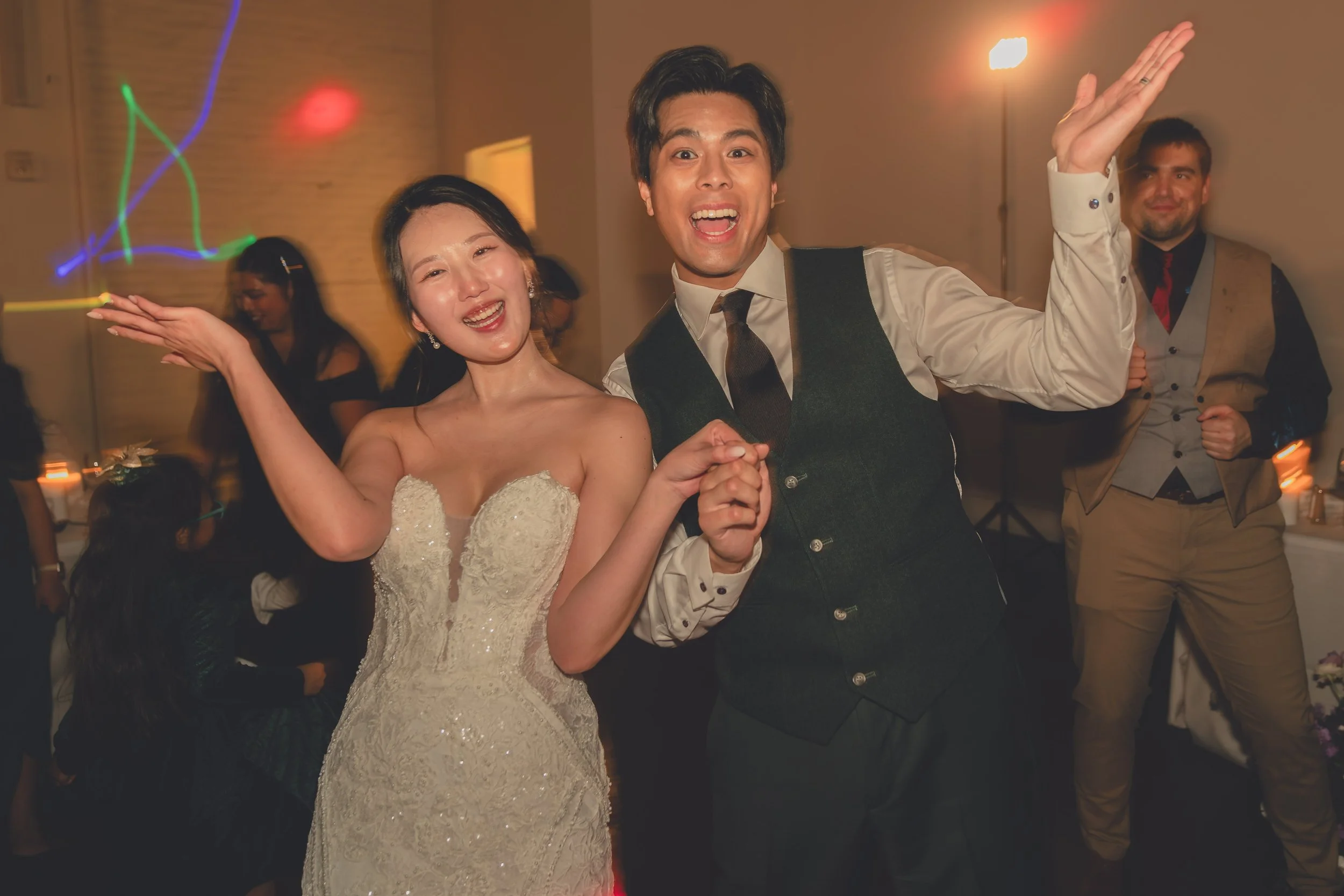 Young couple dancing at a wedding reception, smiling and posing for the camera, with guests and colorful lights in the background.