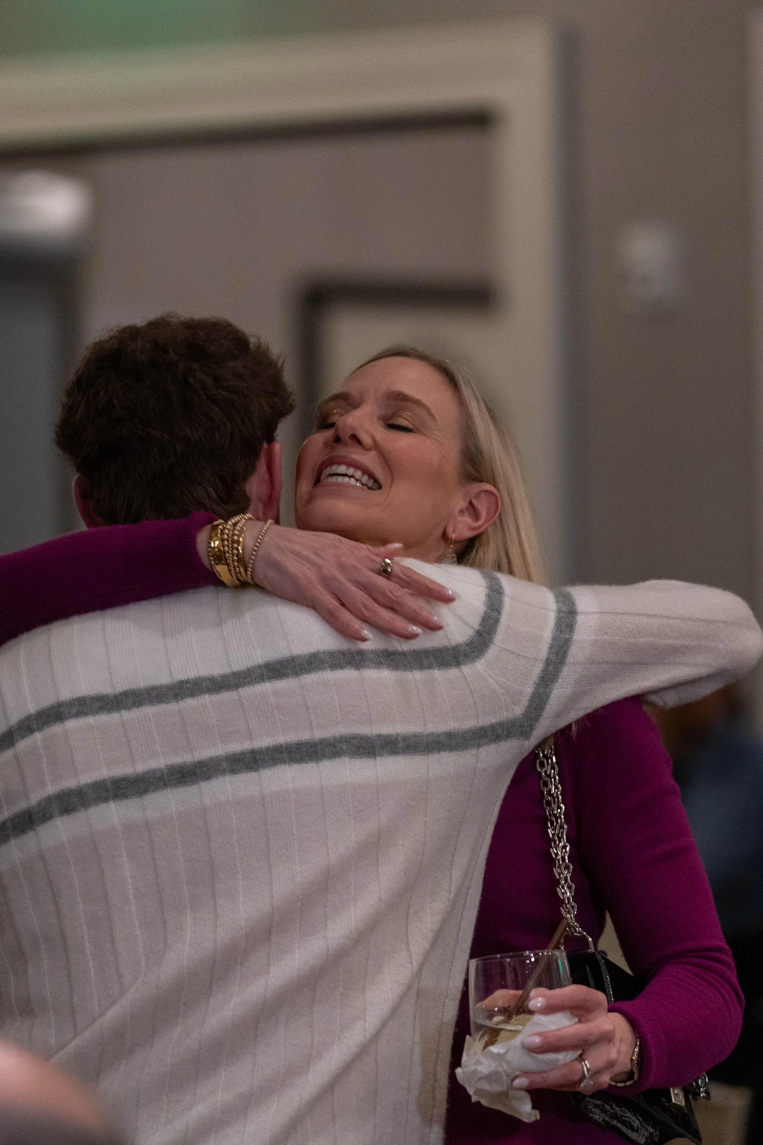 A woman in a purple sweater hugging a man in a striped sweater, smiling, holding a drink with a straw and tissue in her hand.