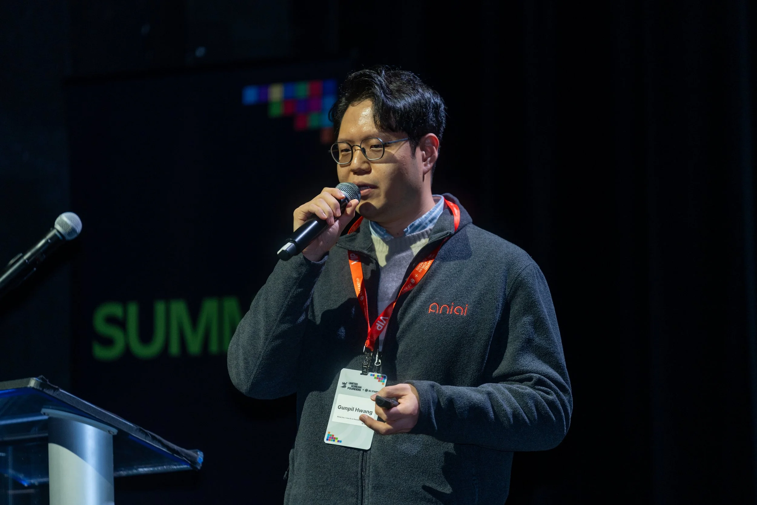 Person speaking at conference with microphone, wearing a gray sweater and red lanyard, standing near a podium with a second microphone.