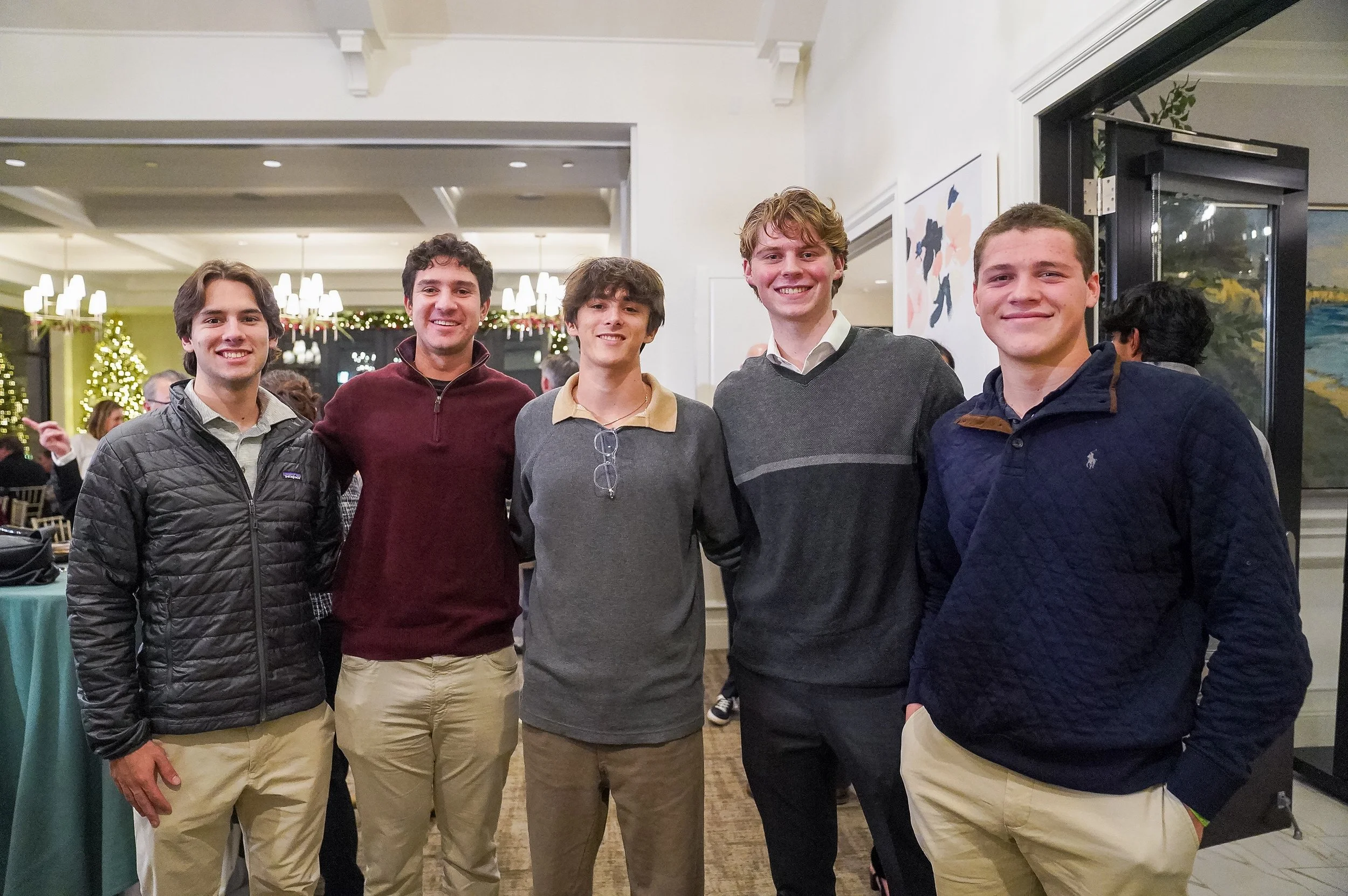 Five young men standing together indoors, dressed in casual attire, with festive decorations in the background.