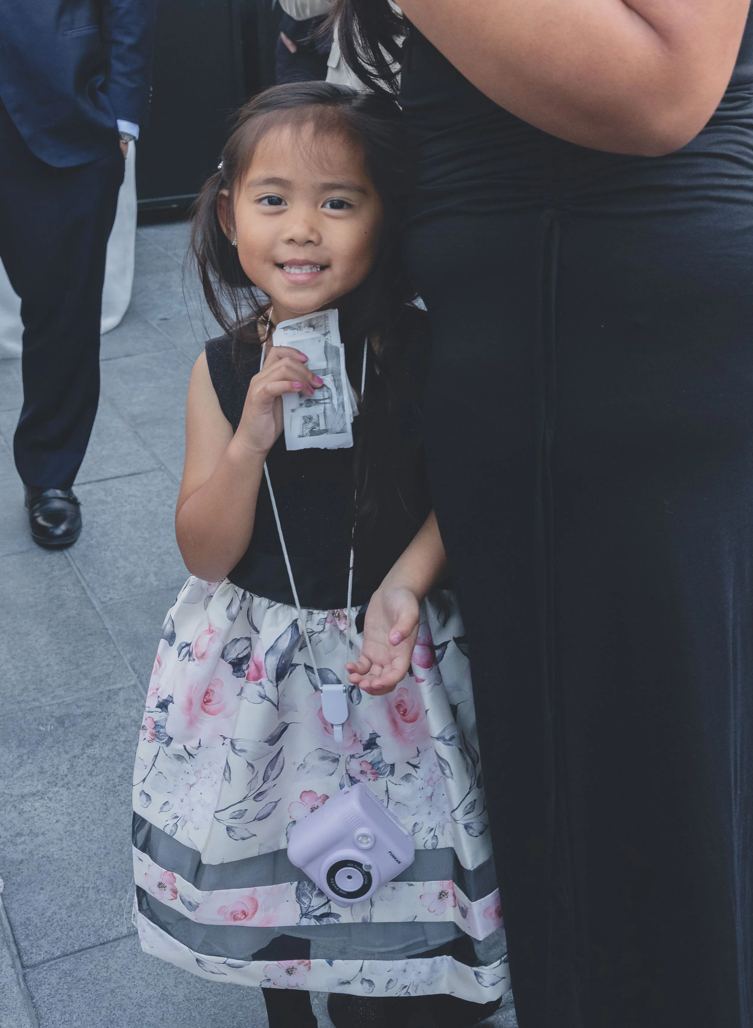 Young girl in floral dress holding instant photos, smiling at a formal event.