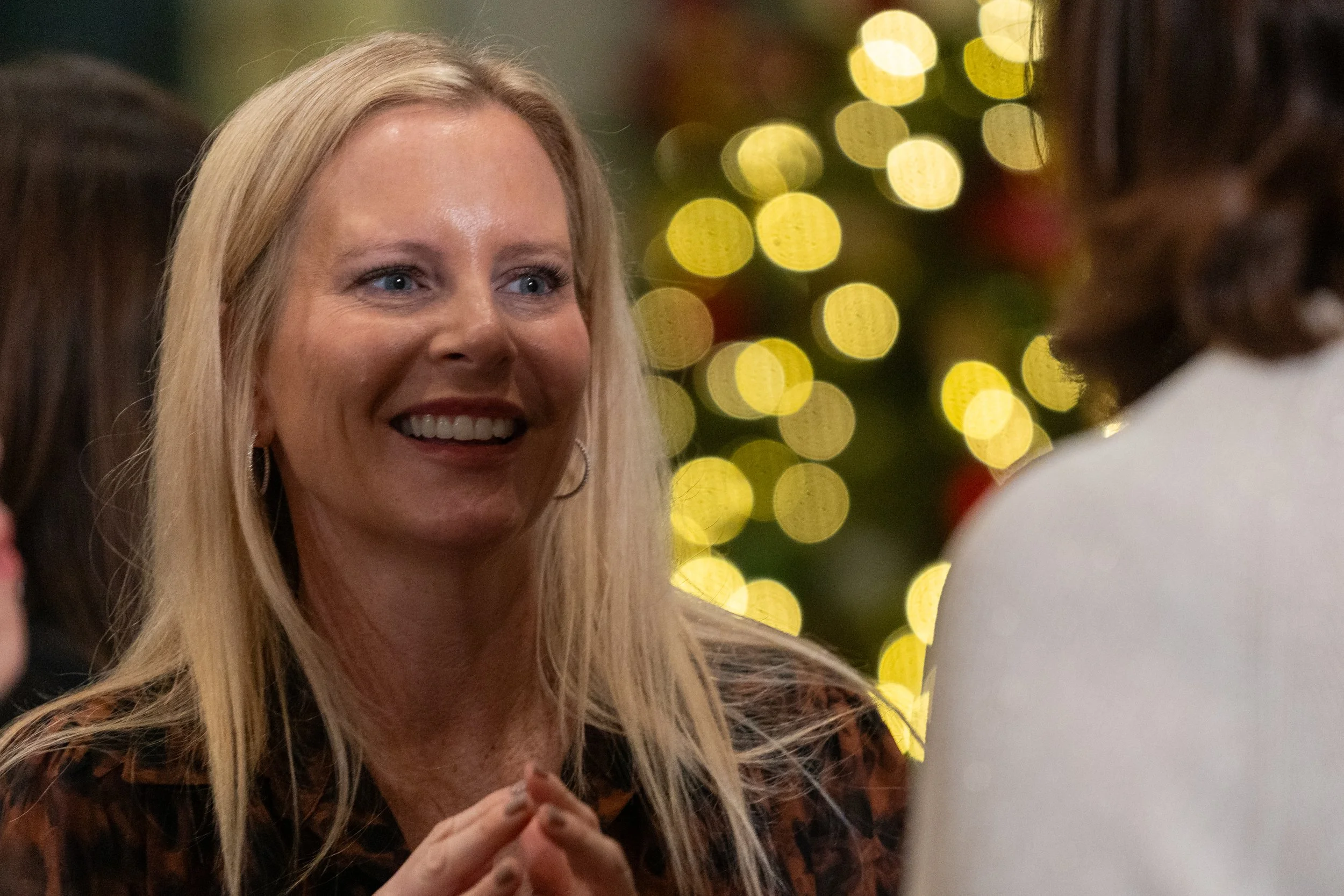 Smiling woman with blonde hair at a social gathering with bokeh lights in the background, engaging in conversation.