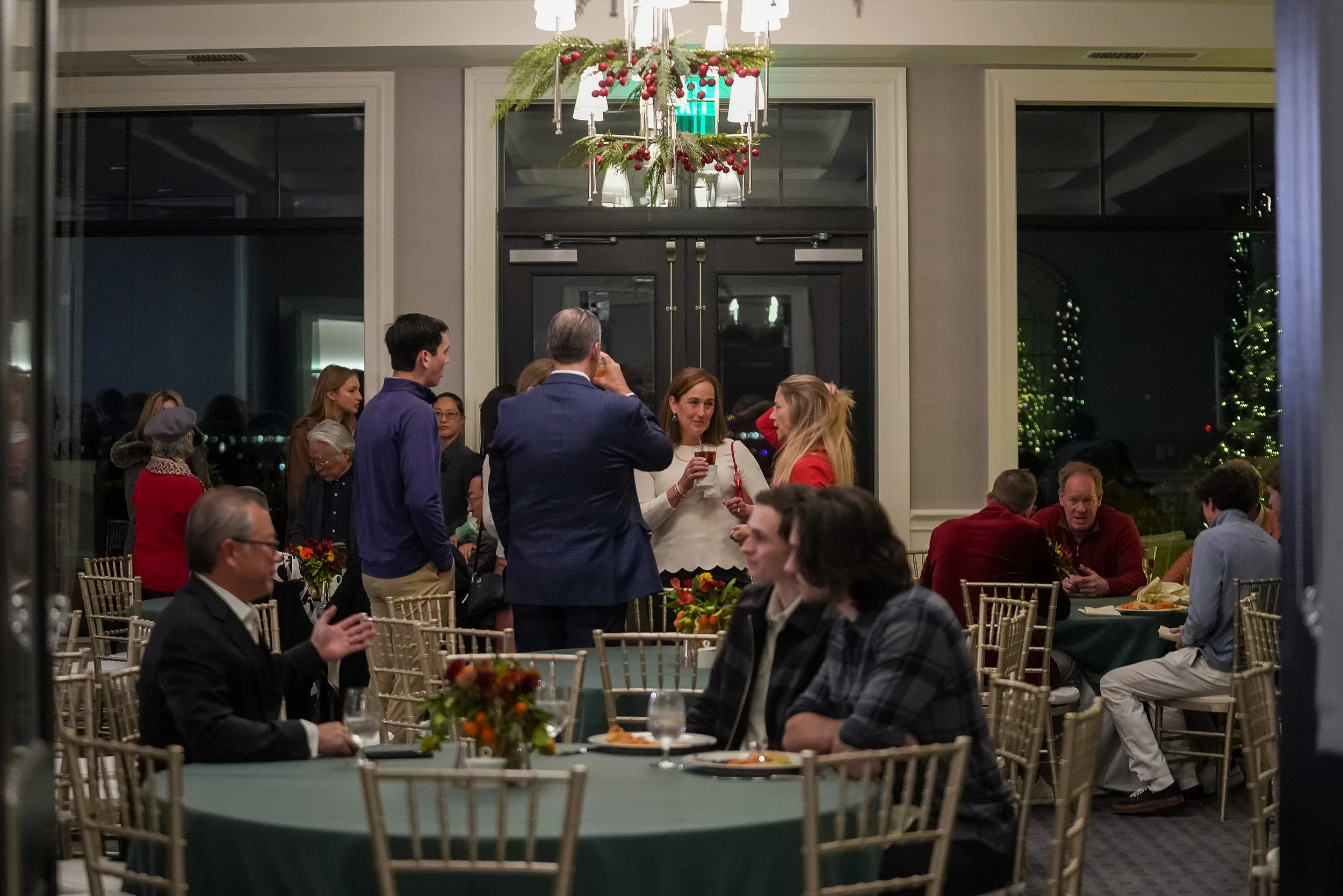 People at a social gathering indoors, standing and sitting around tables. The room is decorated with holiday-themed decor, including plants on tables and a chandelier with ornaments. Attendees are dressed in semi-formal attire, engaging in conversati
