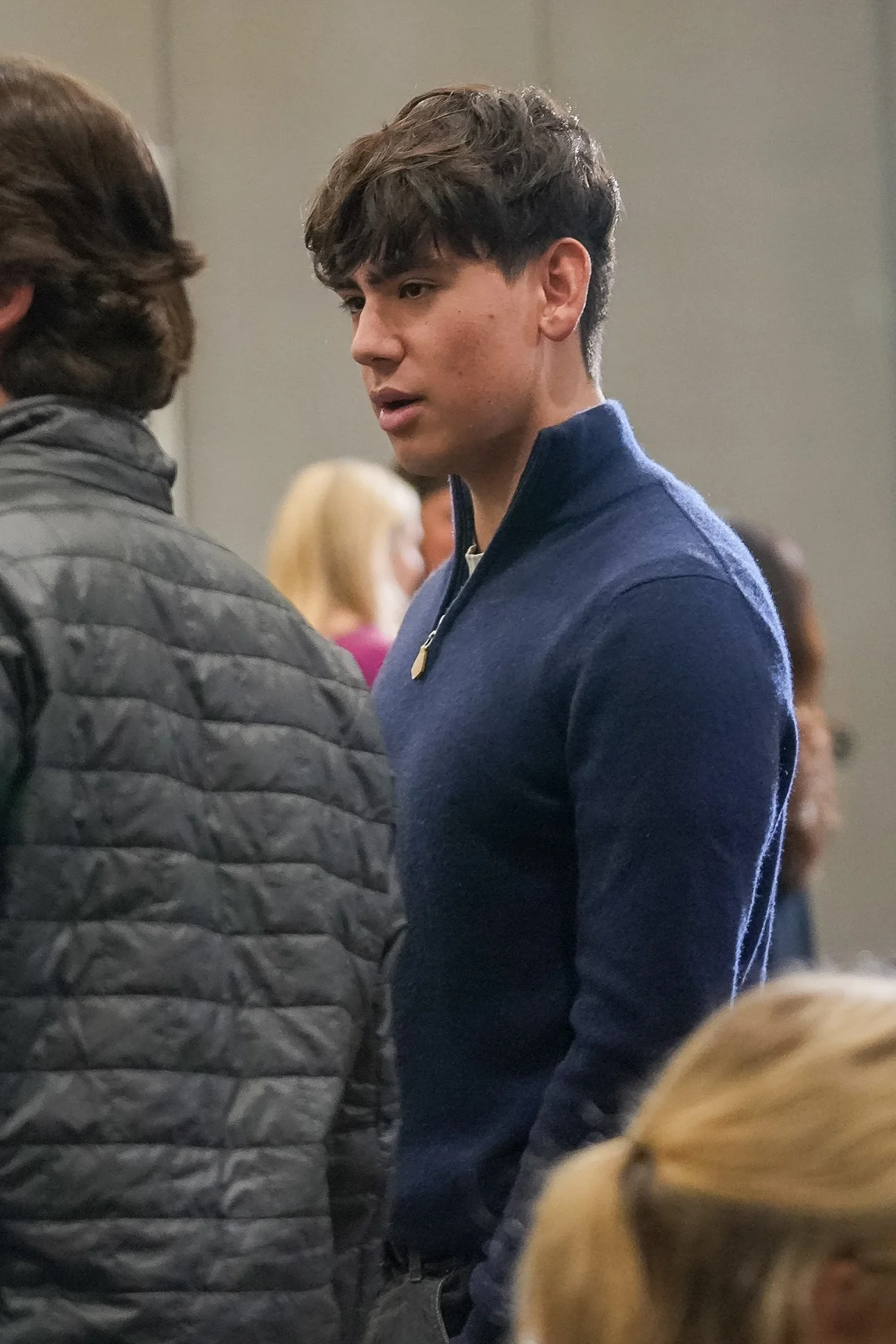 A man in a blue sweater standing among people indoors, possibly at an event.