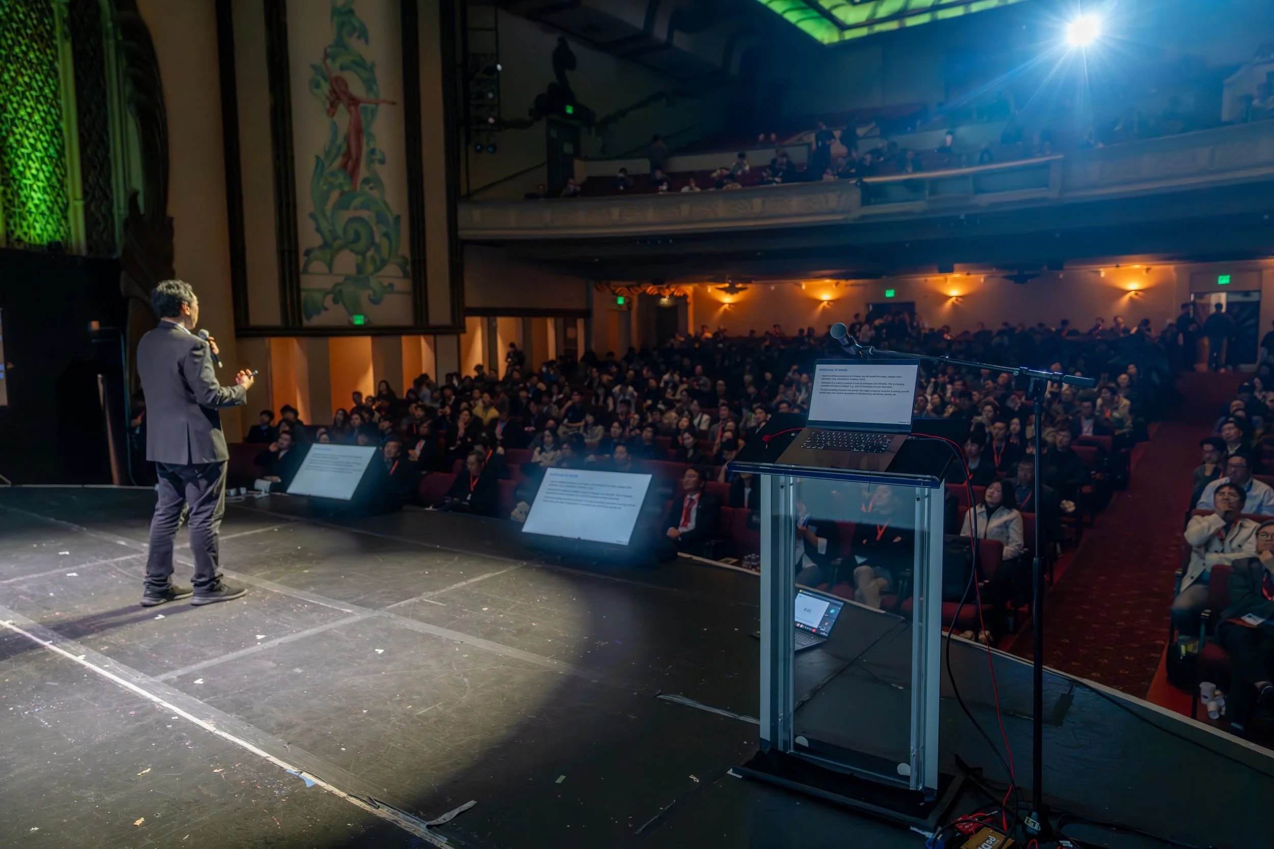 A speaker on stage addressing a large audience in a theater setting. The podium with a microphone and a laptop is visible, and the audience is seated in rows, attentively listening. The venue features ornate decorations and a balcony with additional