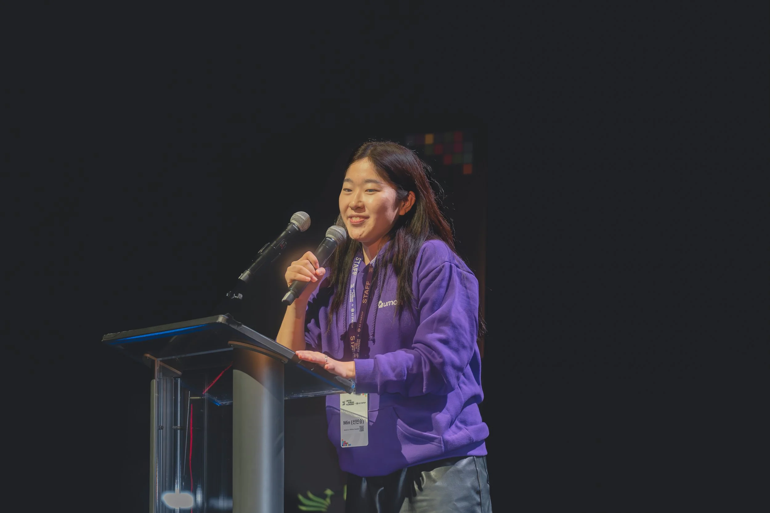 Person speaking at a podium with a microphone, wearing a purple hoodie and lanyard, against a dark background.