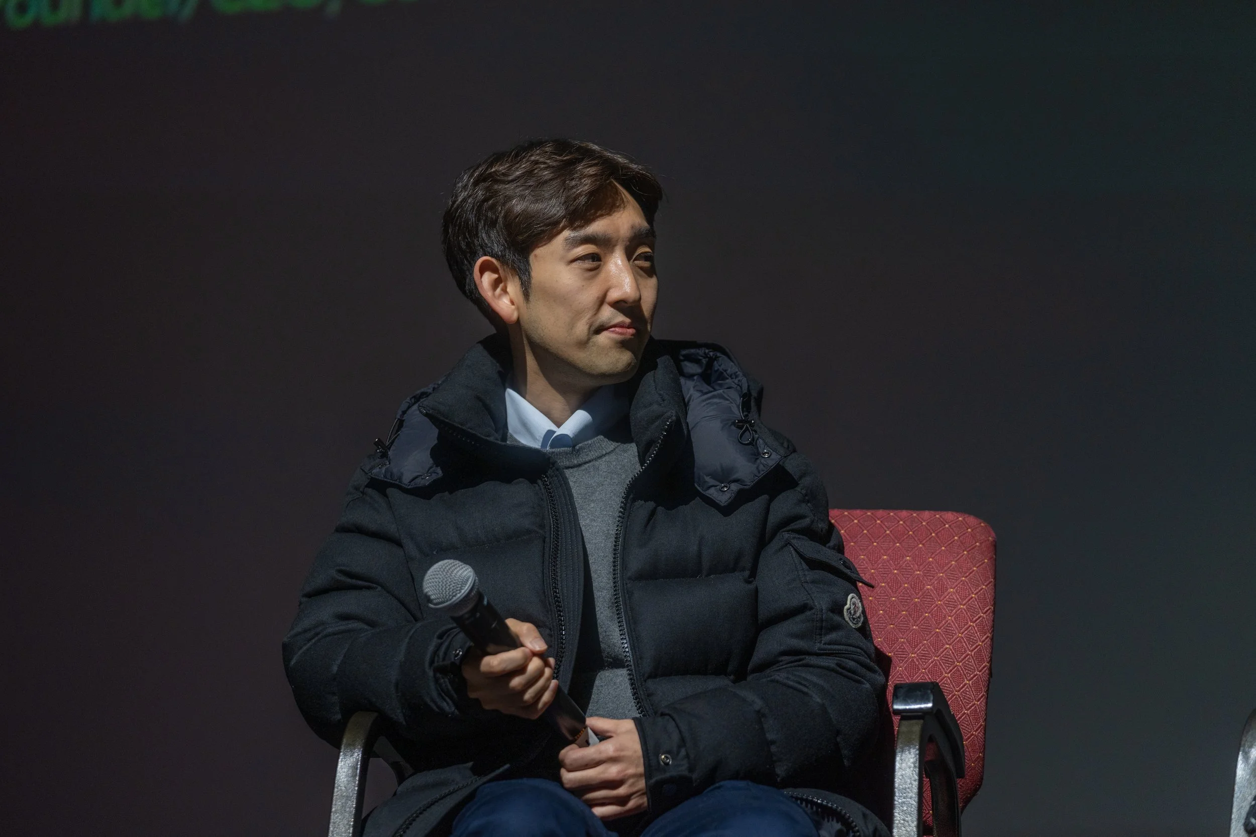 A person sitting on a chair holding a microphone, wearing a black coat and looking to the side, against a dark background.