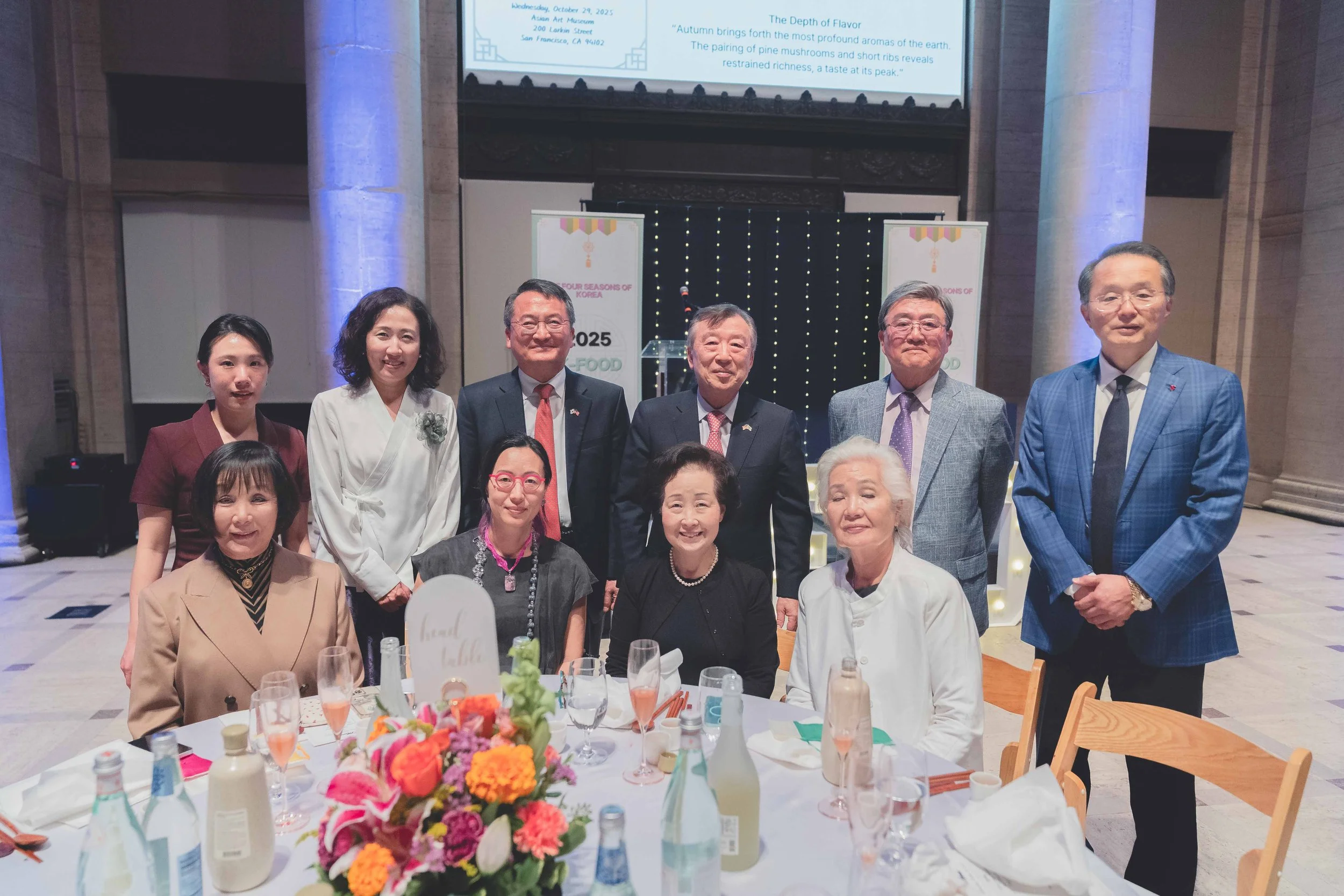 Group of eleven people, including elderly and middle-aged adults, gathered around a table at a formal event, with a large floral centerpiece and drinks. They are indoors, in front of a screen and banners with text.