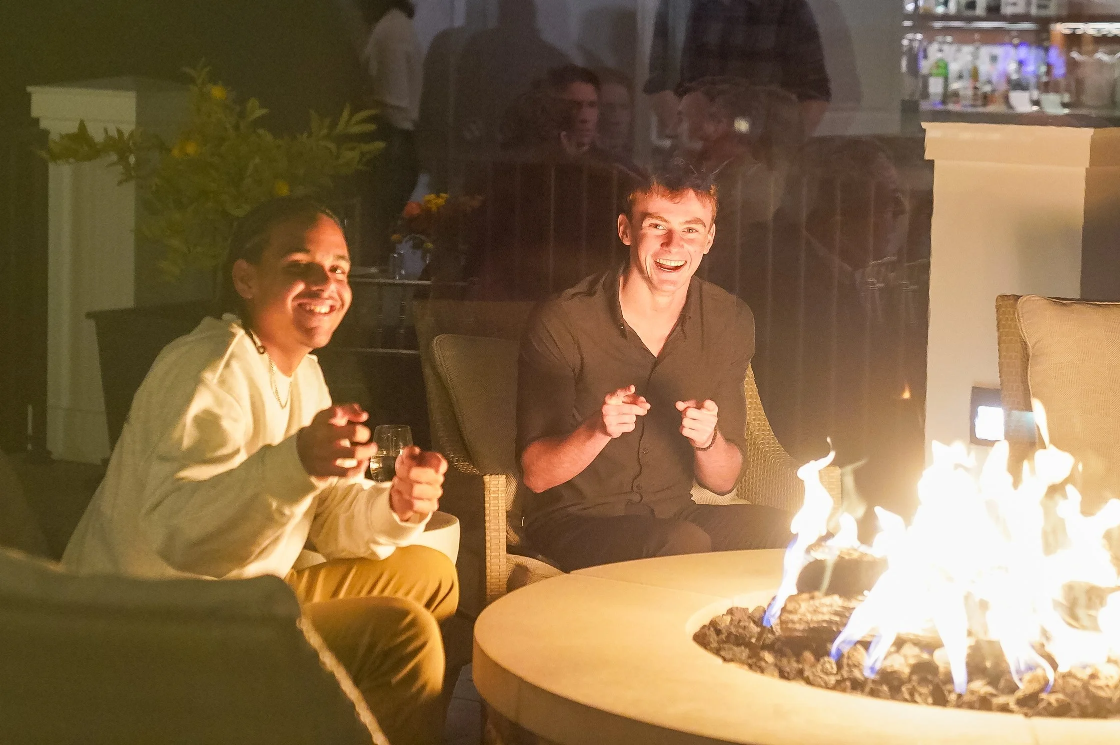 Two people sitting by a fire pit, smiling and pointing, in an outdoor setting with a warm ambiance.