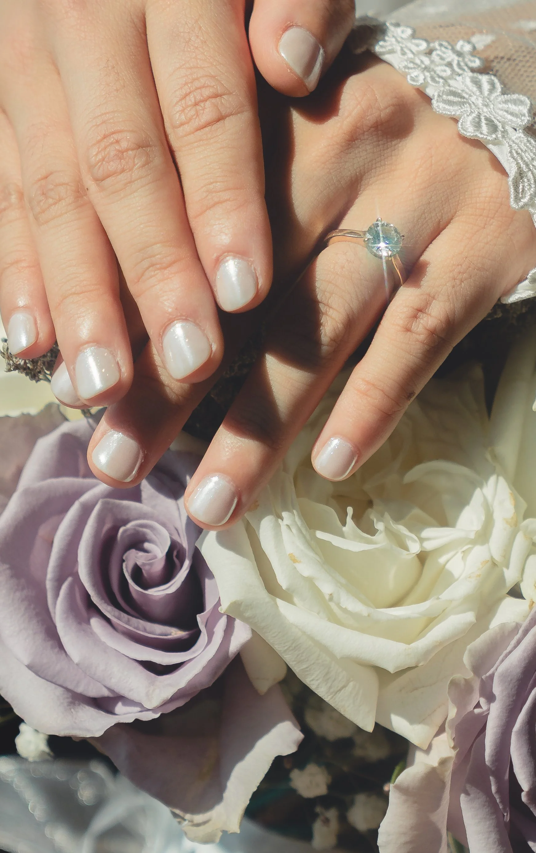 Close-up of a person's hands with manicured nails and a ring with a large gemstone, resting on white and purple roses.