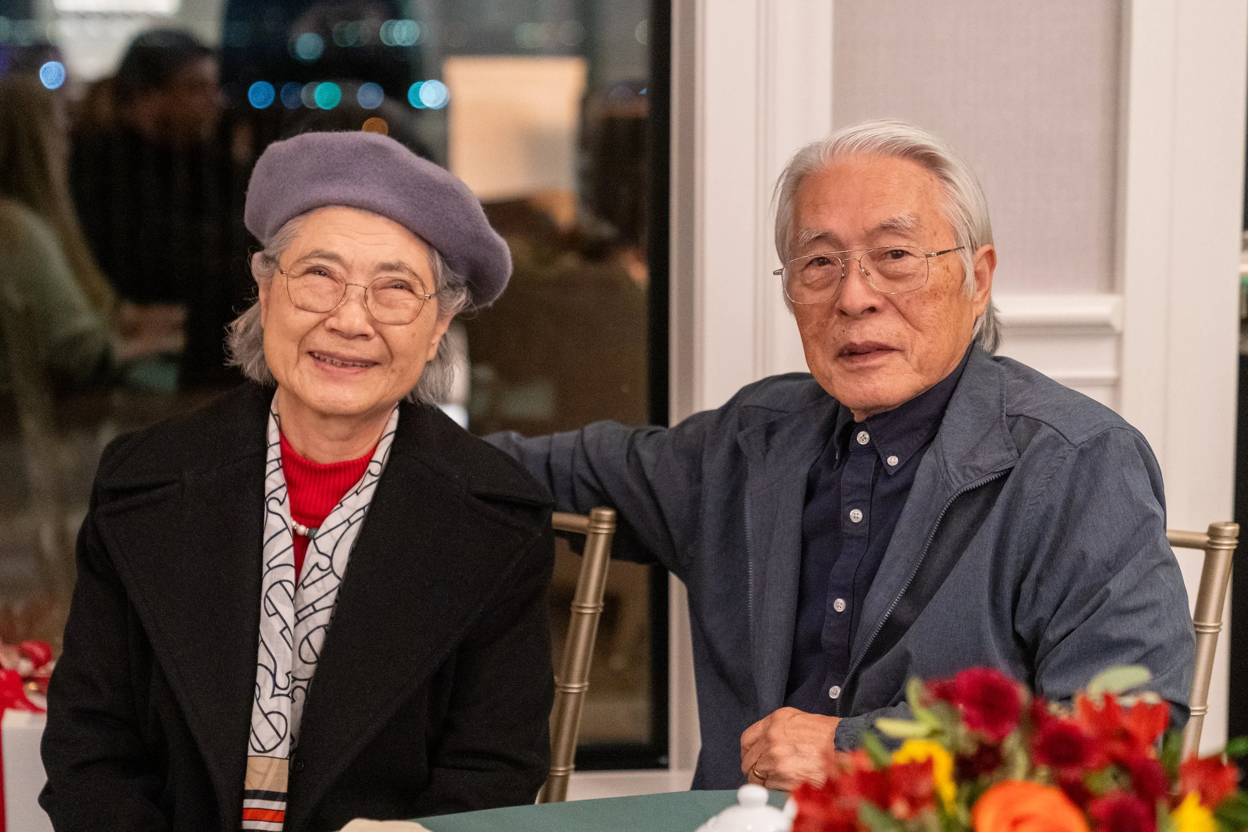 An elderly couple seated together at a table indoors, with the woman wearing a purple beret and glasses, and the man wearing glasses and a dark jacket. A floral arrangement is on the table in the foreground.
