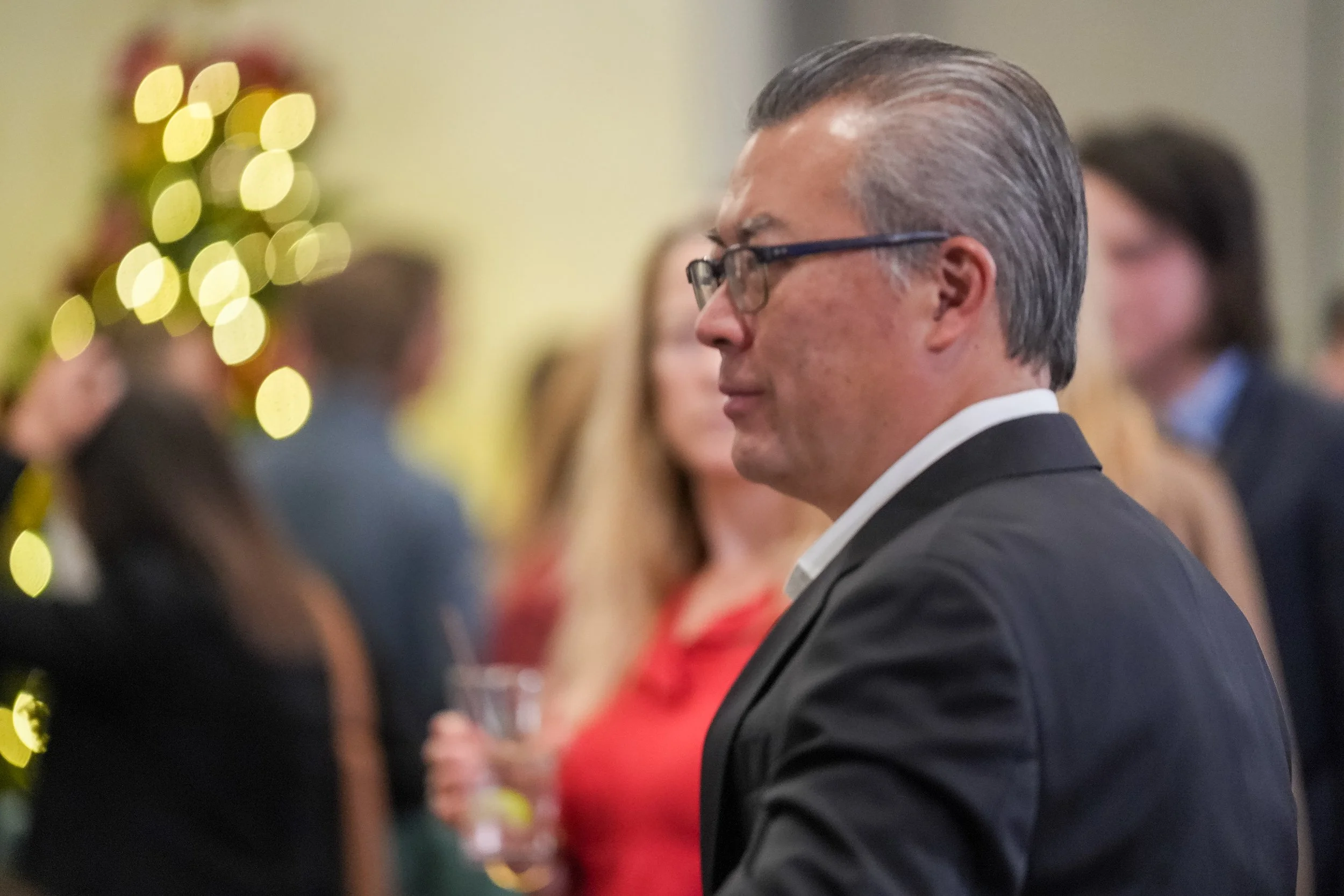 A man in glasses and a suit at a social event with blurred people in the background and festive lights.
