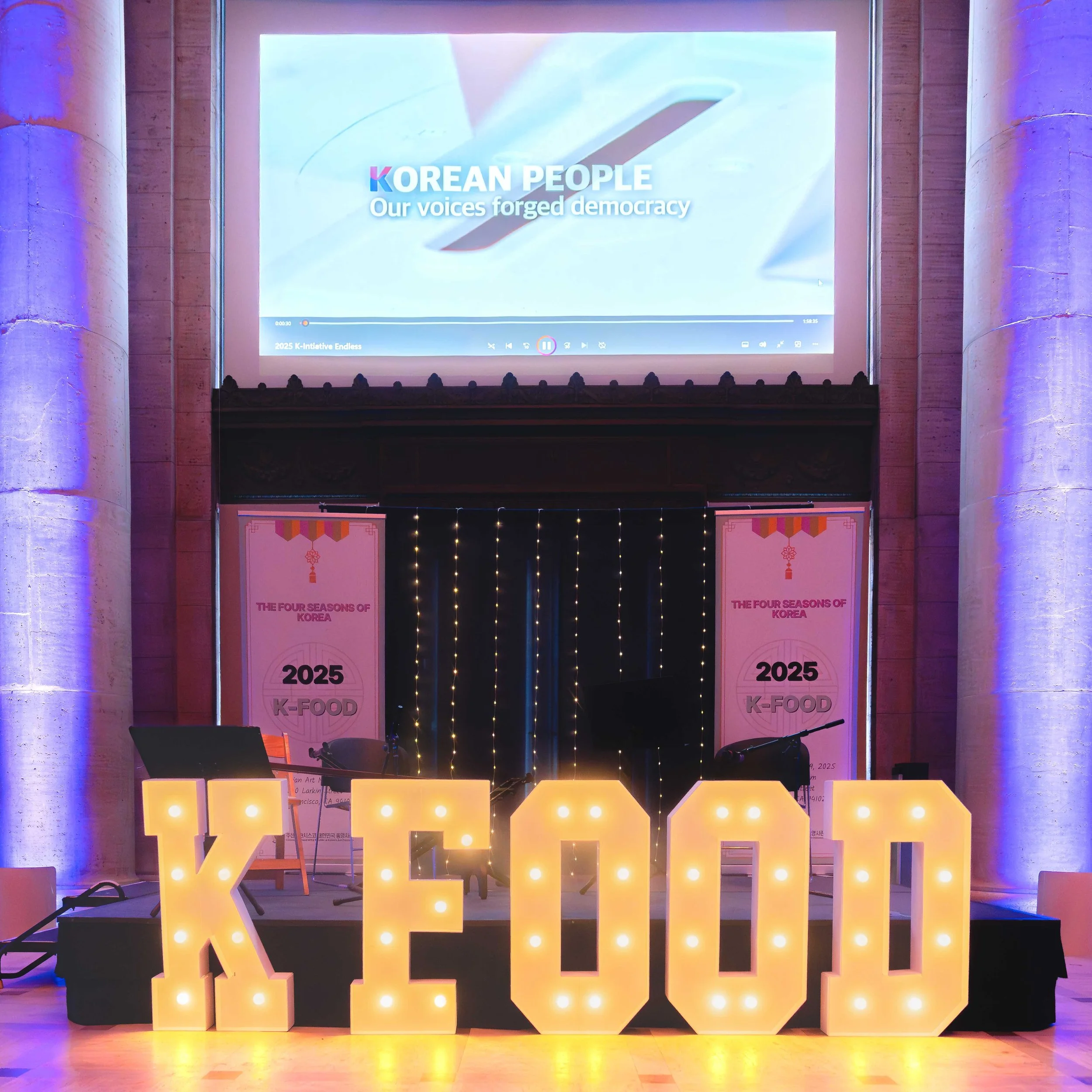 Stage decorated with large illuminated letters spelling KEFOOD, with banners behind reading 'The Four Seasons of Korea' and '2025 K-Food'. A large screen above displays a Korean-themed video, highlighting 'Korean People' and 'Our voices forged democr