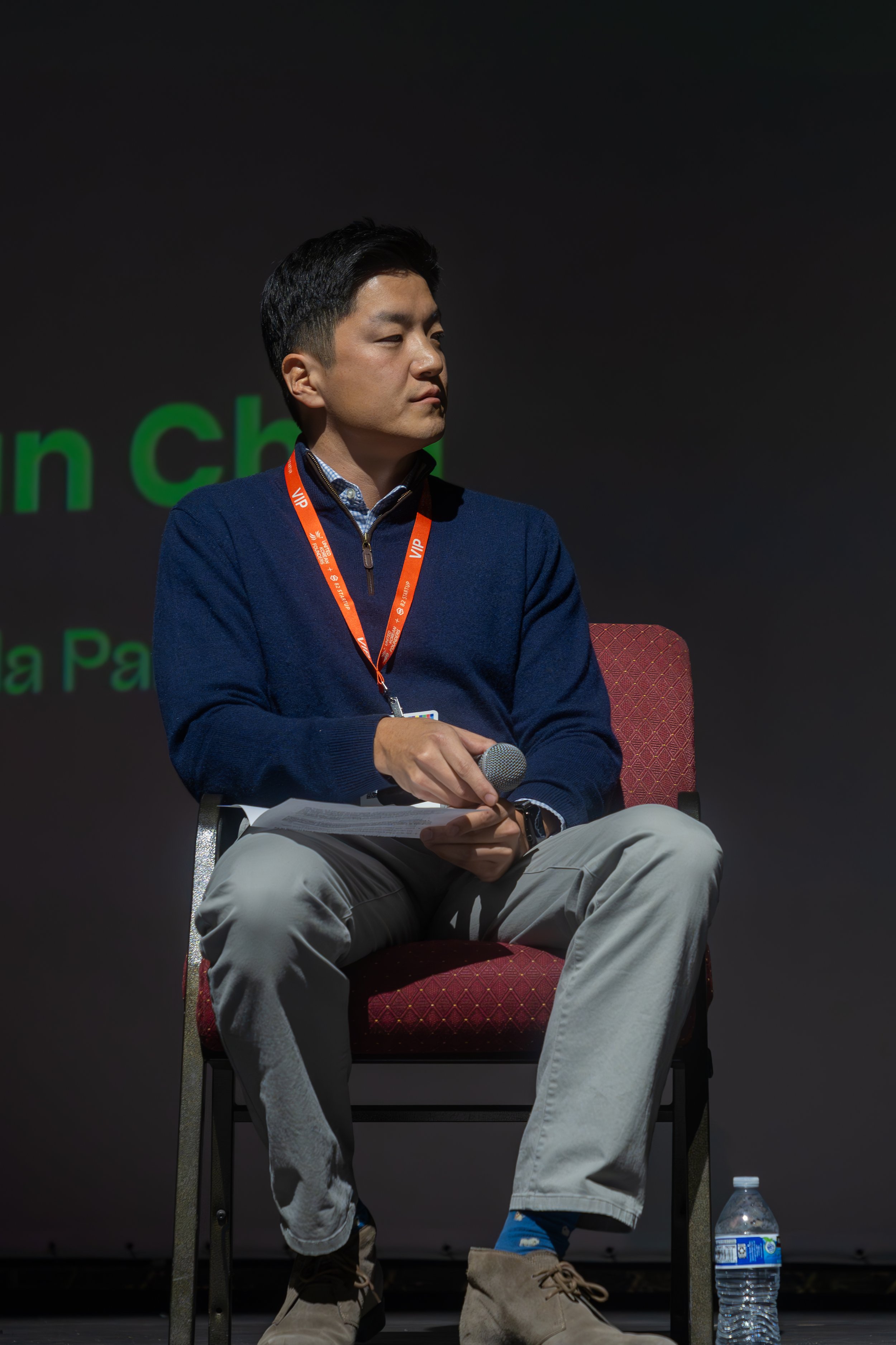 Man seated on stage holding a microphone, wearing a blue sweater and orange lanyard, with a water bottle on the floor beside him.