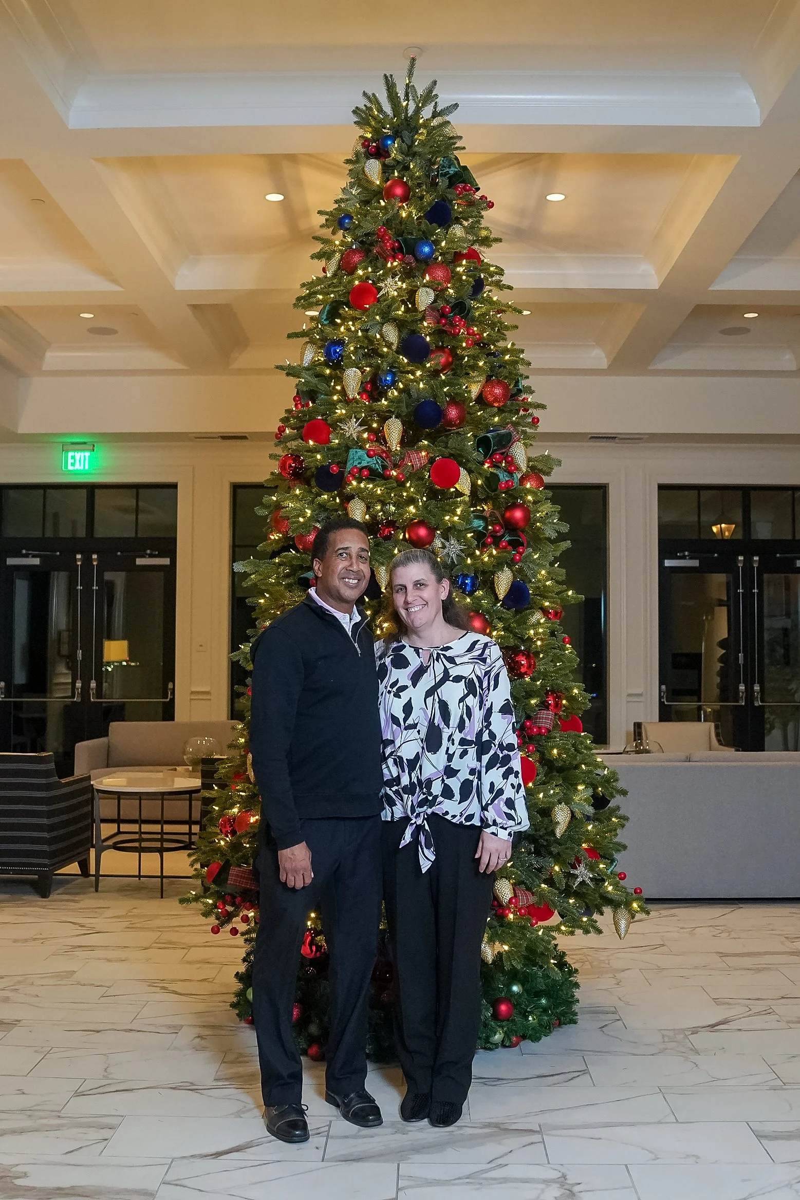 A couple posing in front of a large decorated Christmas tree indoors.