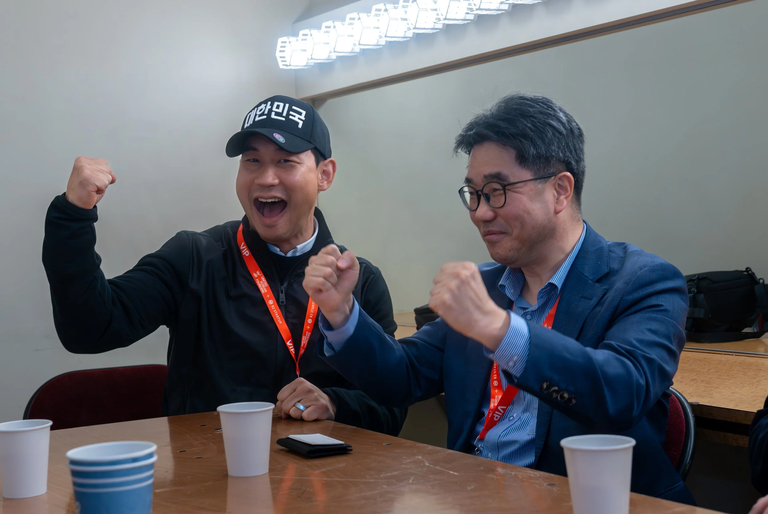Two men celebrating at a table with paper cups, wearing lanyards and showing excitement with raised fists.