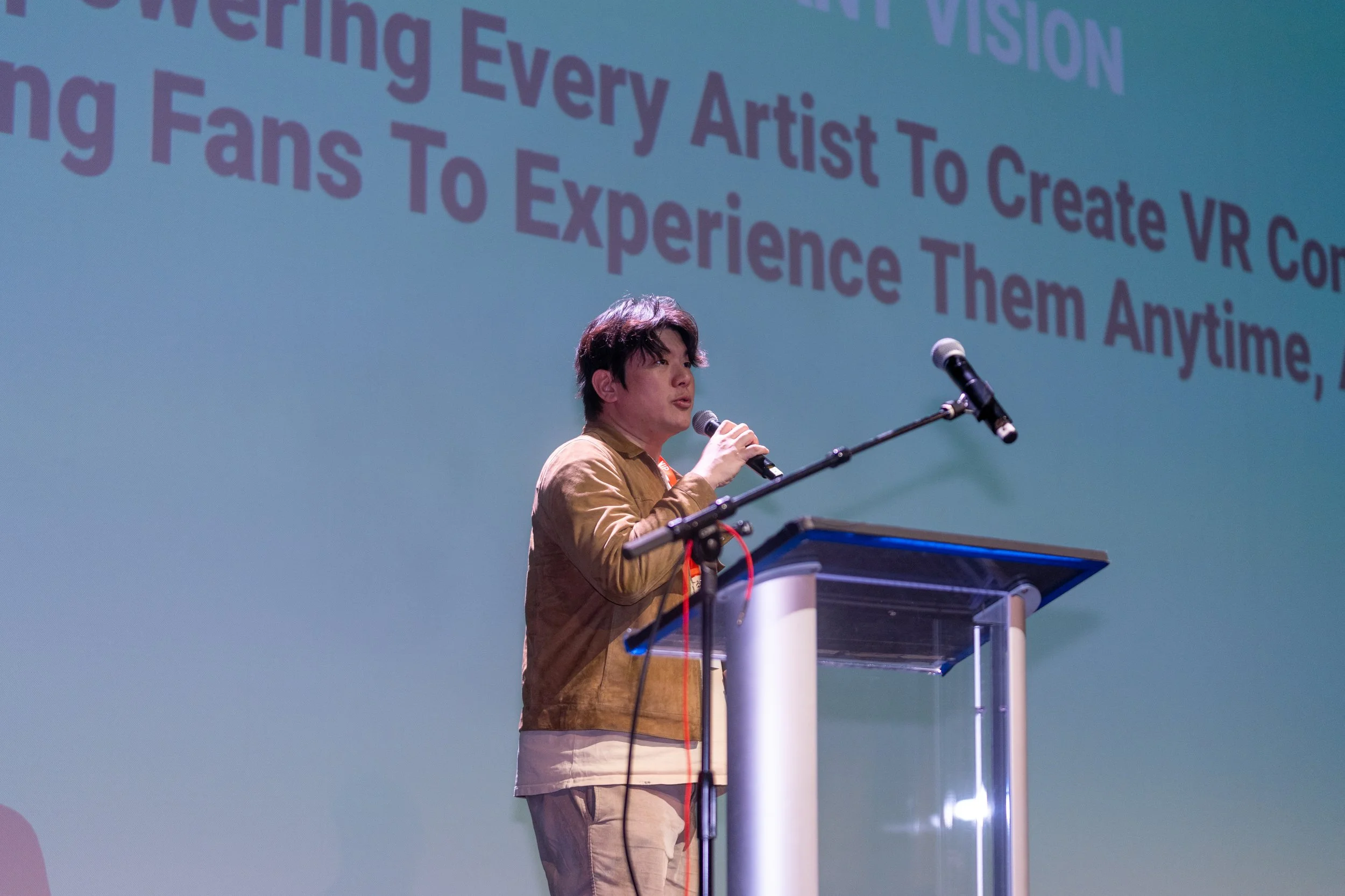 Person speaking at a podium with a microphone, in front of a projection screen displaying a partial text about VR content and fans.