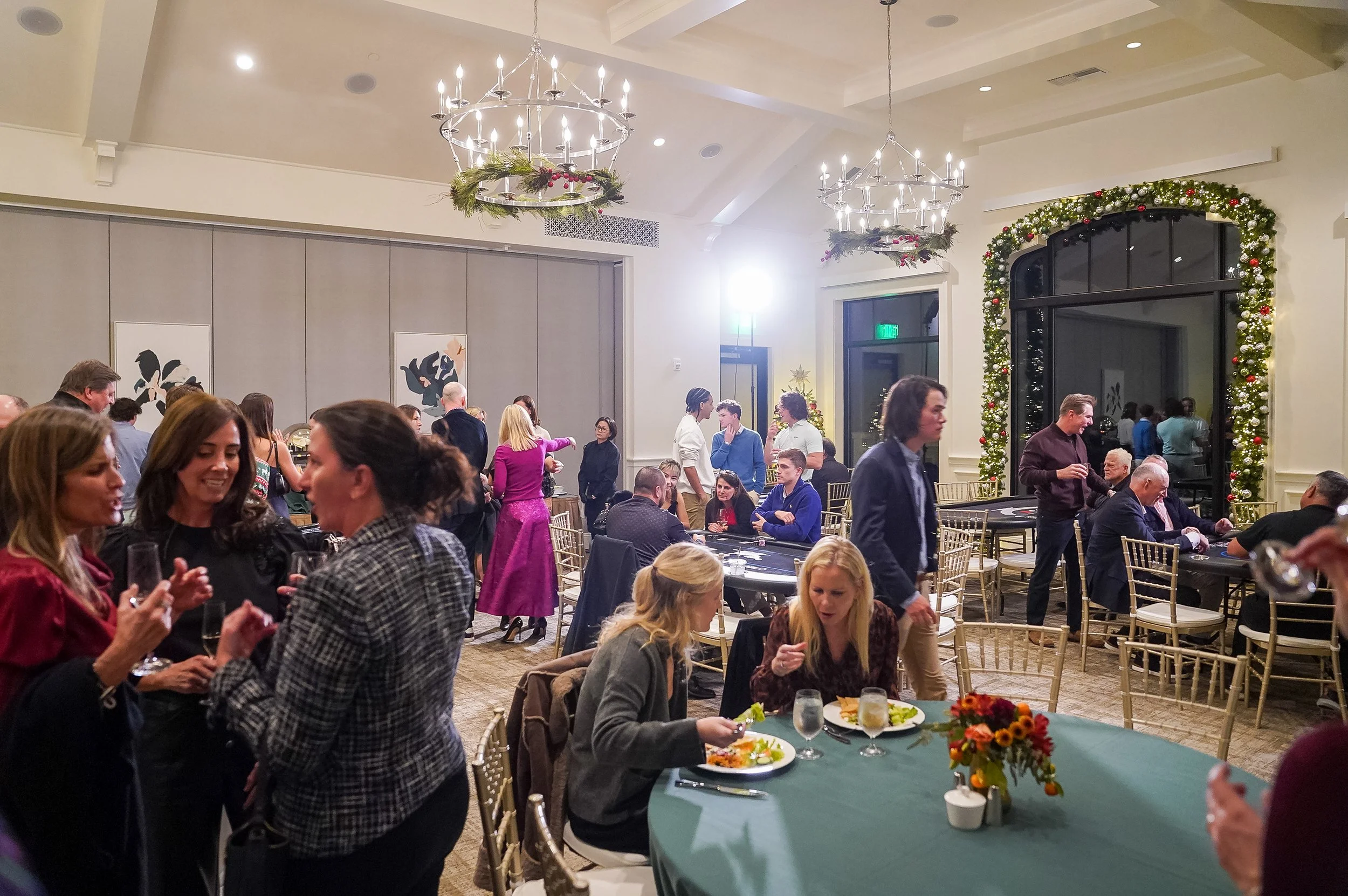 People socializing and dining in a festive event hall with holiday decorations and chandeliers.