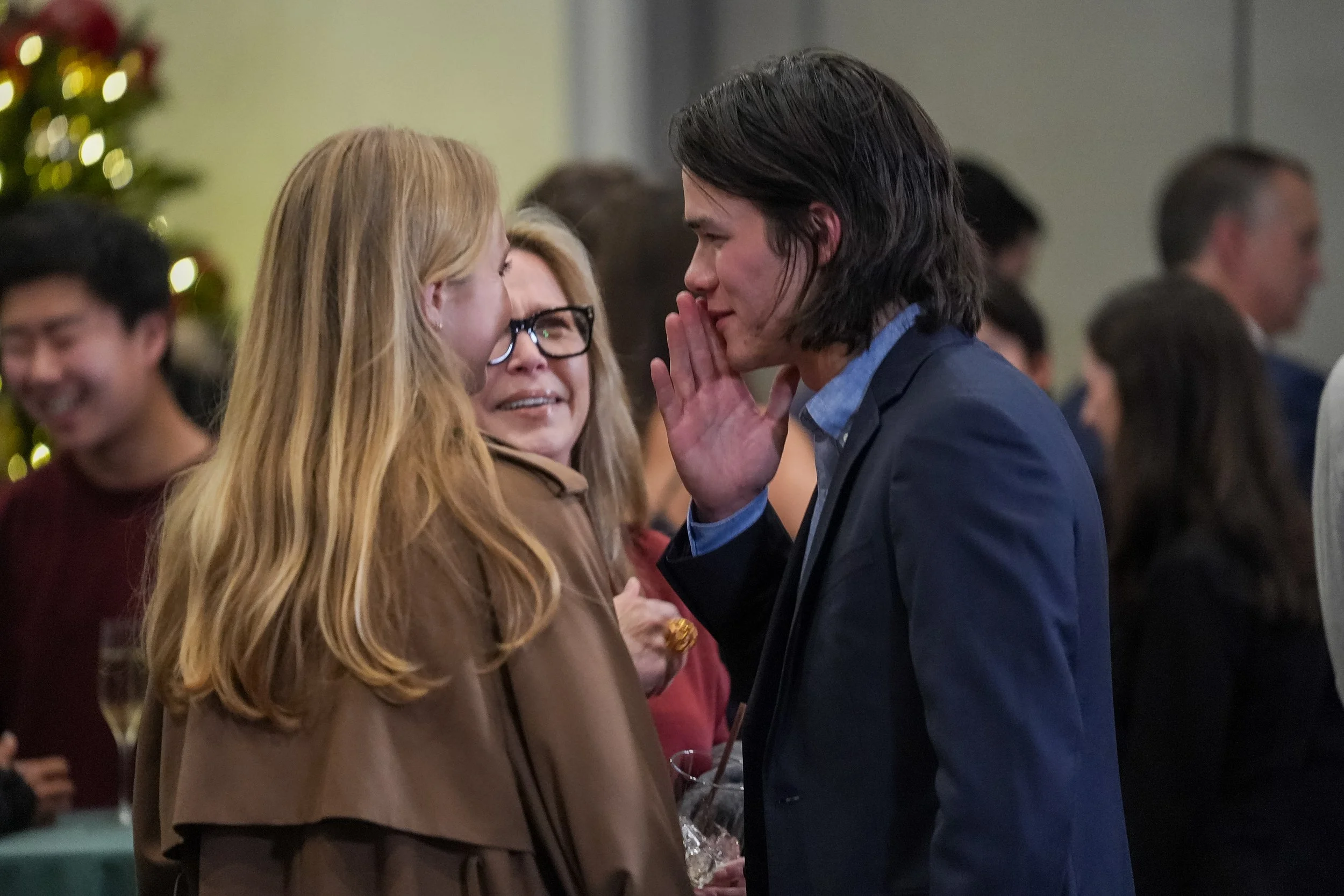 Group of people at a social gathering, one man whispering to a woman in a crowded room.