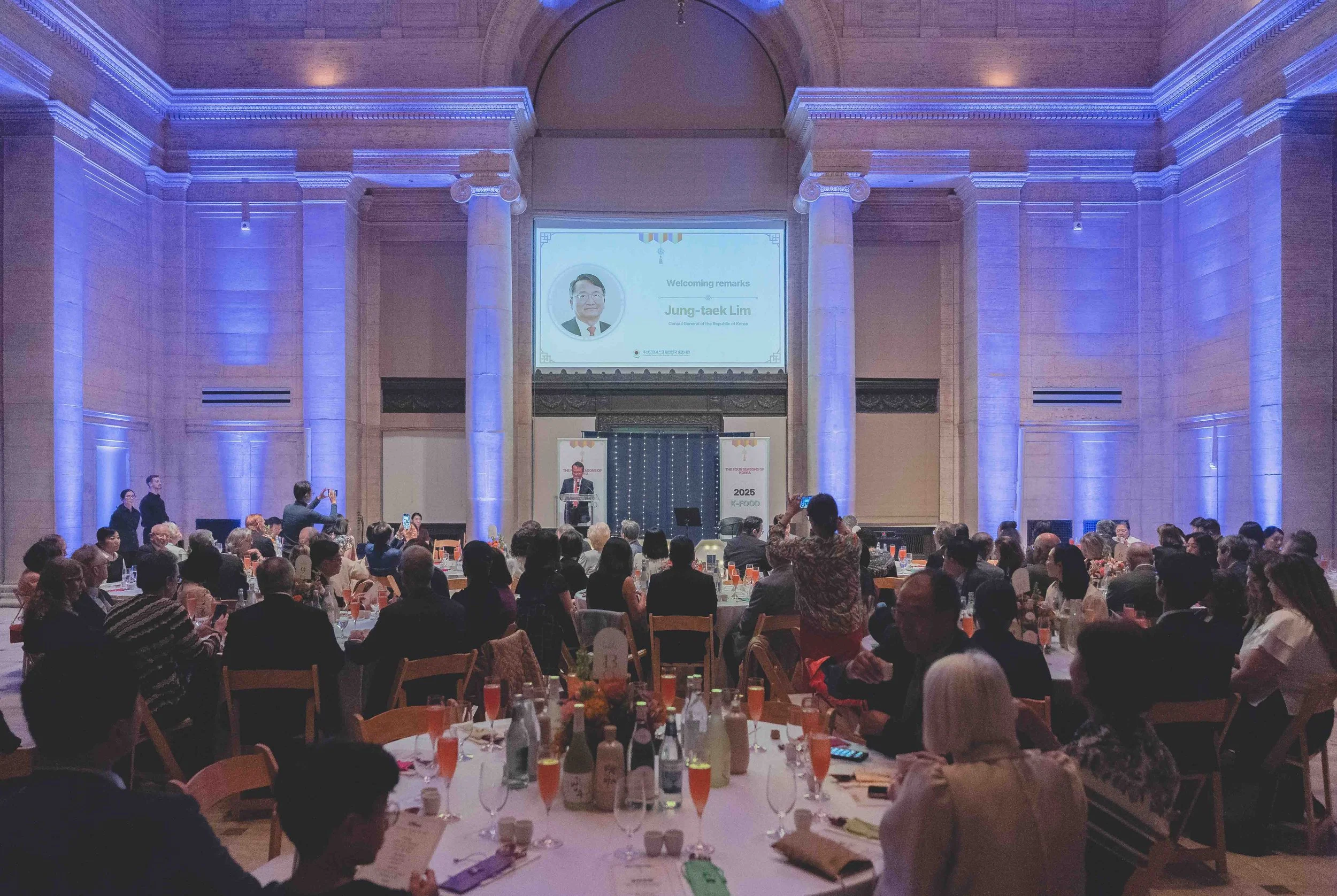 A large formal event with many attendees seated at round tables, facing a stage where a speaker is giving a presentation. The room features high ceilings, decorative columns, and is illuminated with blue lighting.