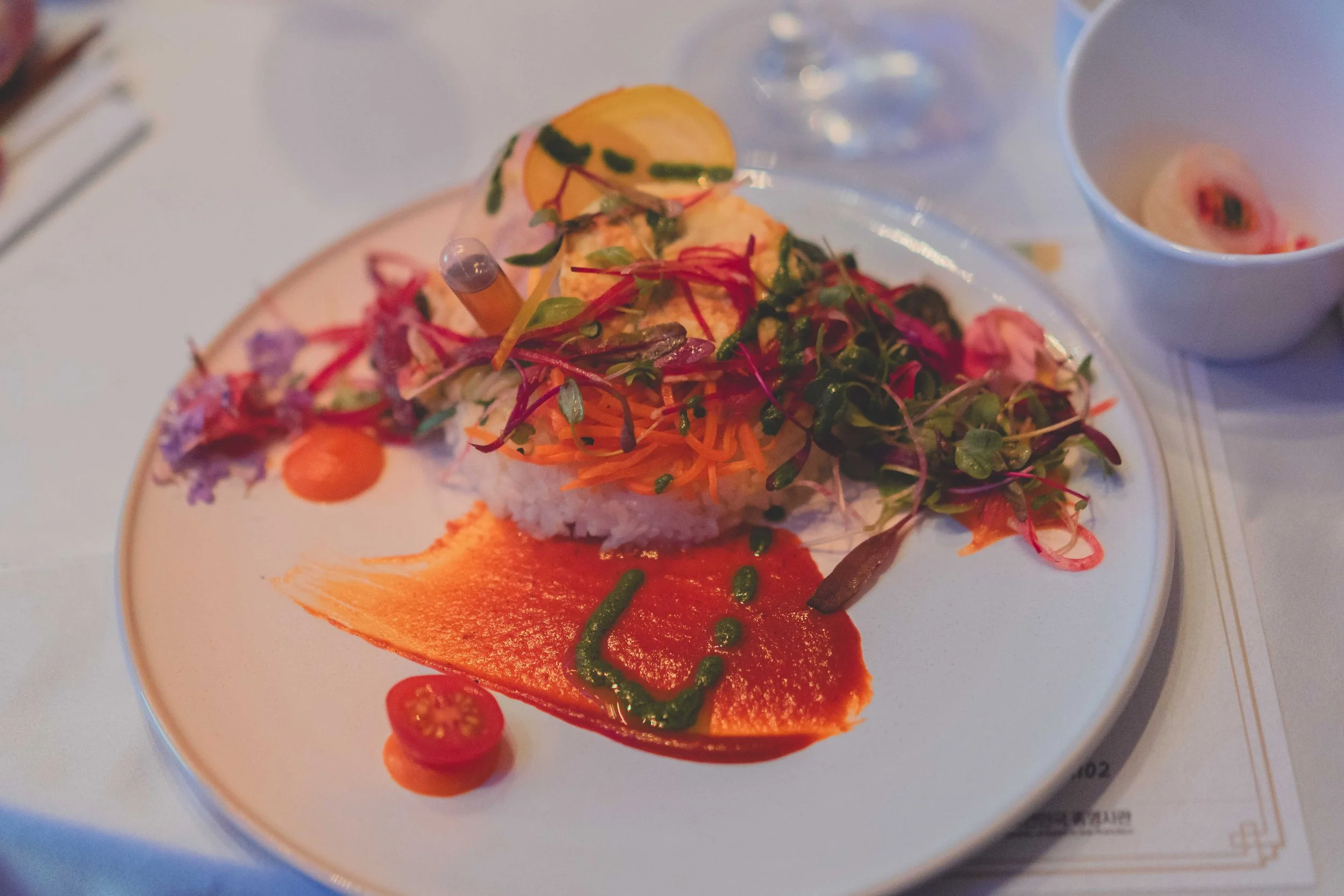 Gourmet plate with rice, salad, carrots, cherry tomatoes, sauce, lemon wedge, and hot dog with vegetables, next to a bowl of dressing on a white table.