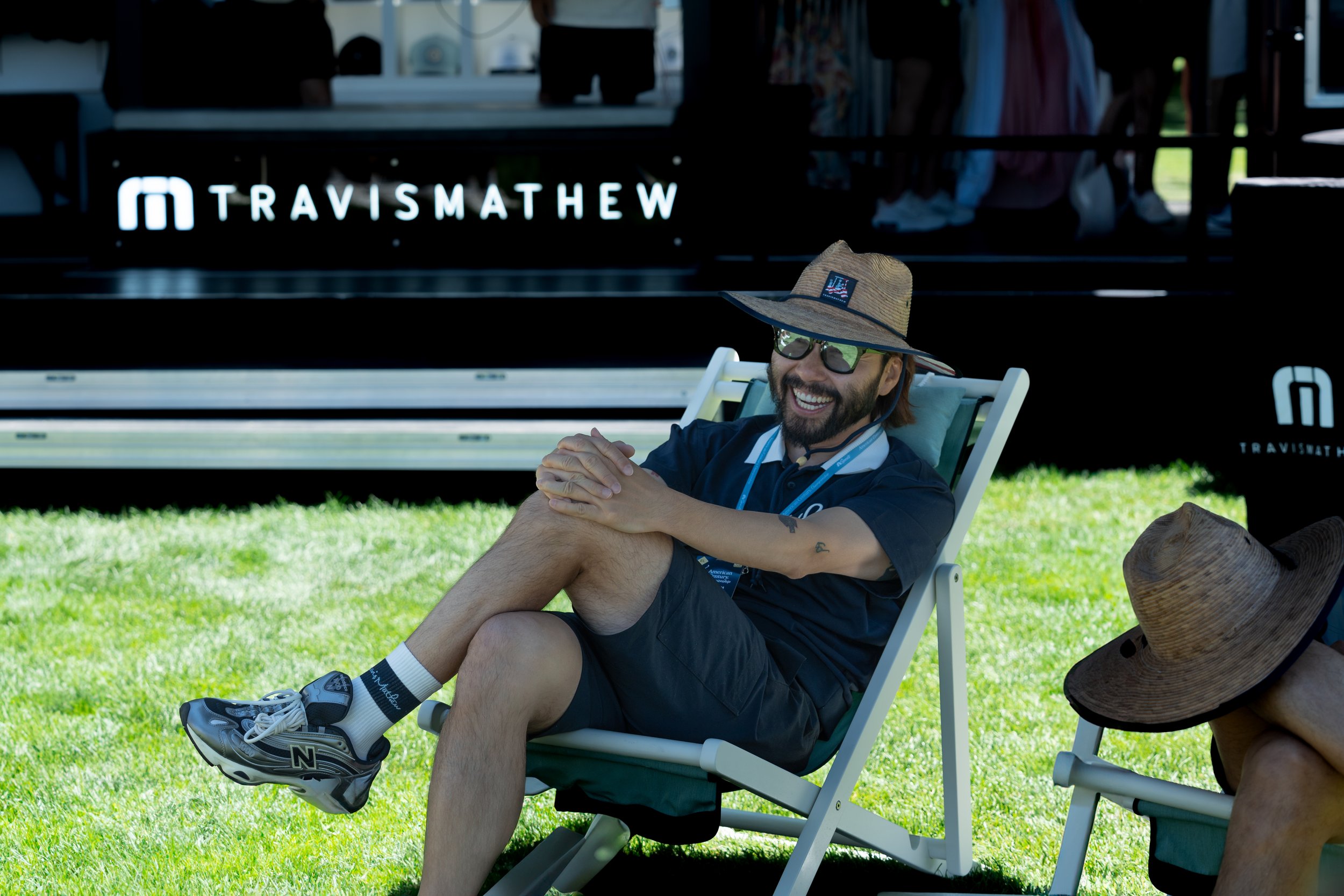 Man smiling and relaxing in a chair on grass, wearing sunglasses and a straw hat, with a "Travis Mathew" sign in the background.