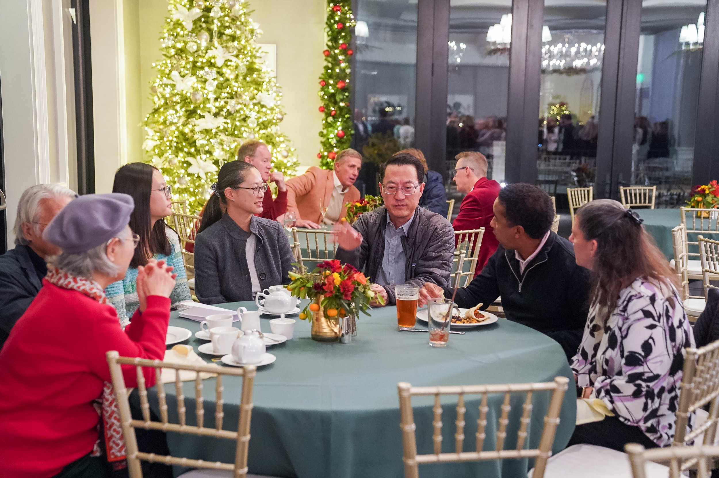 A festive gathering with people seated around a table adorned with a floral centerpiece, enjoying drinks and food. A lit Christmas tree and garland provide a festive backdrop.