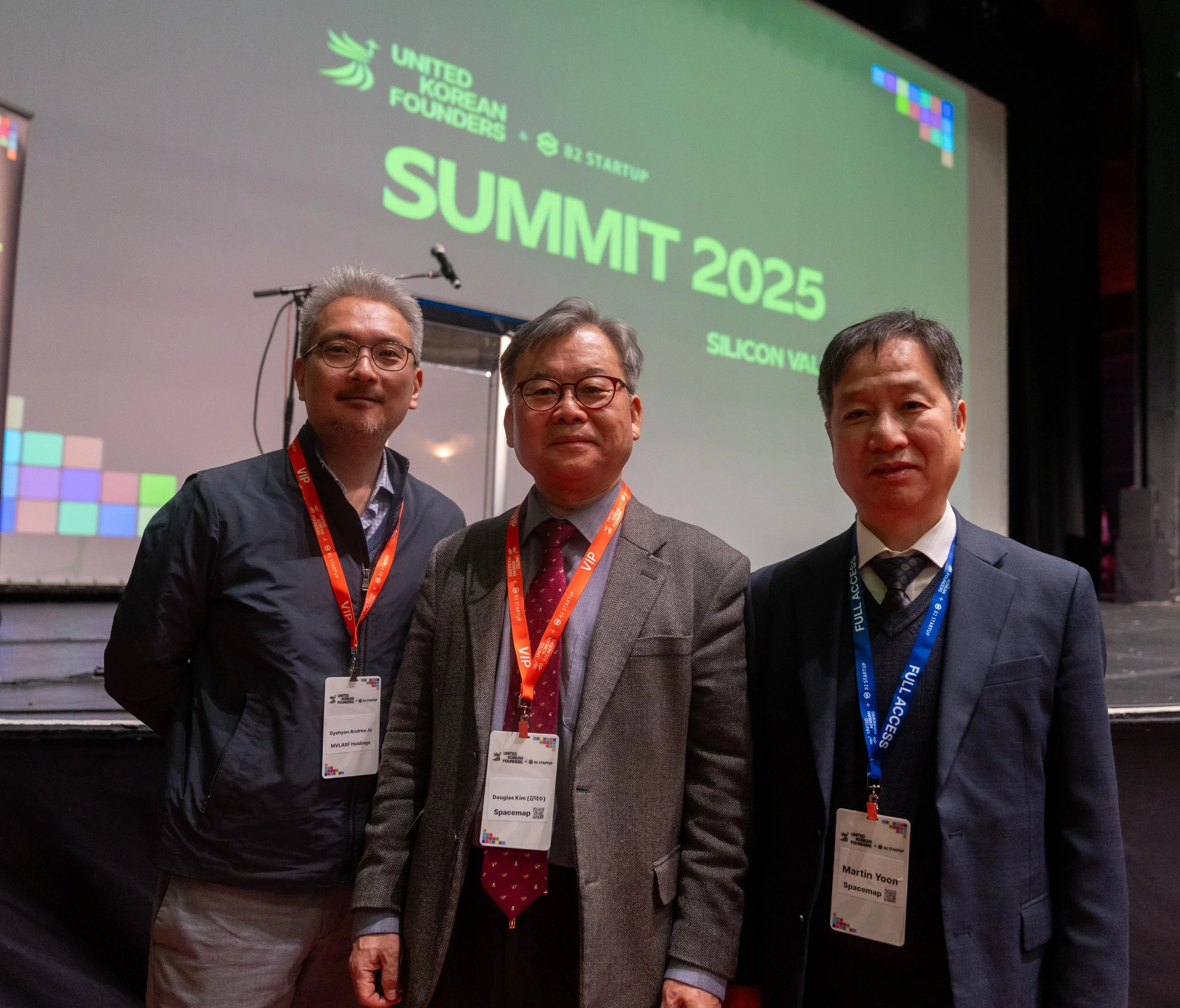 Three men wearing lanyards stand in front of a screen displaying a "Summit 2025" event in Silicon Valley.