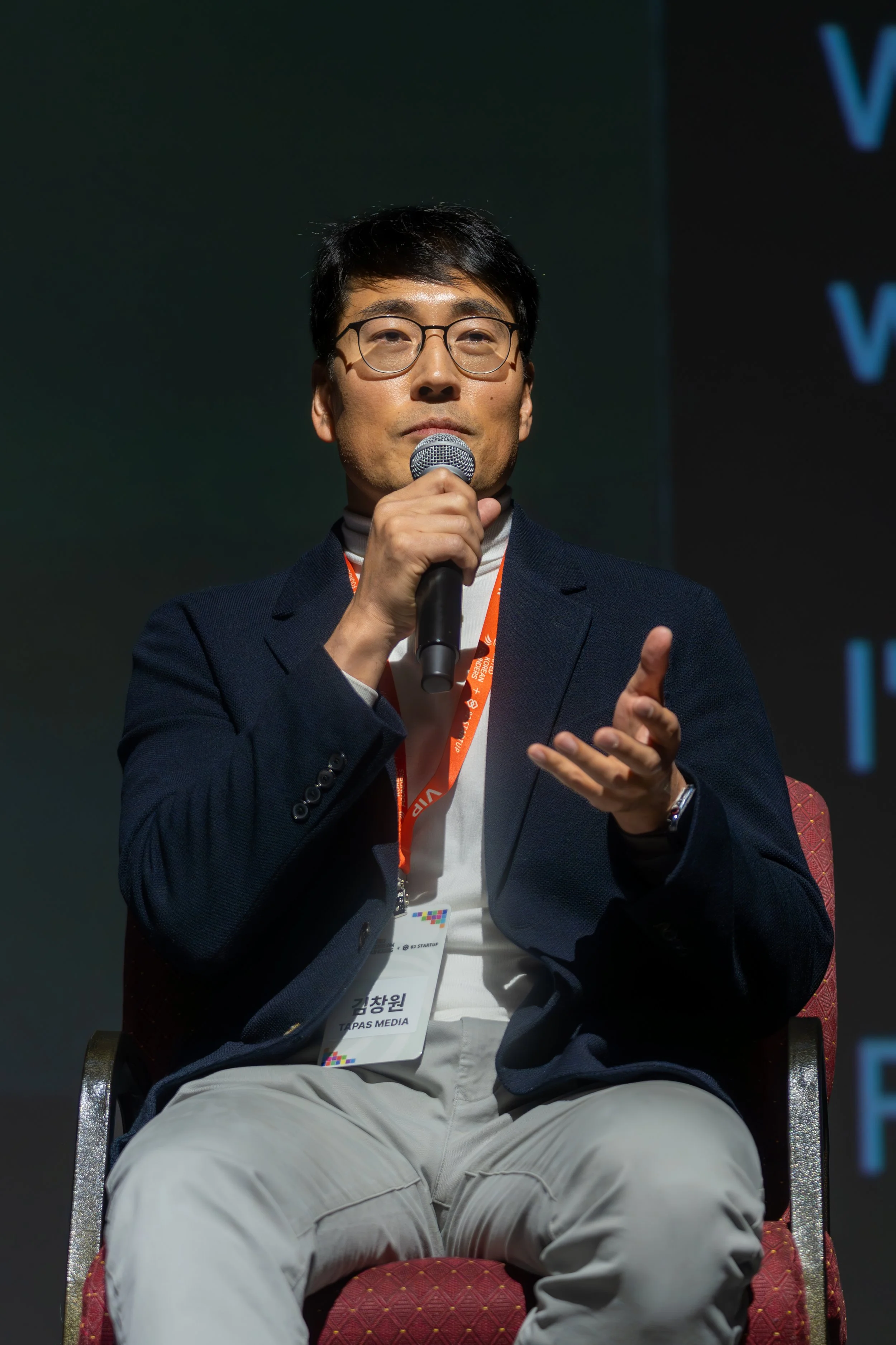 Man in glasses and a suit speaking into a microphone, sitting on a chair, wearing a lanyard with an identification badge.