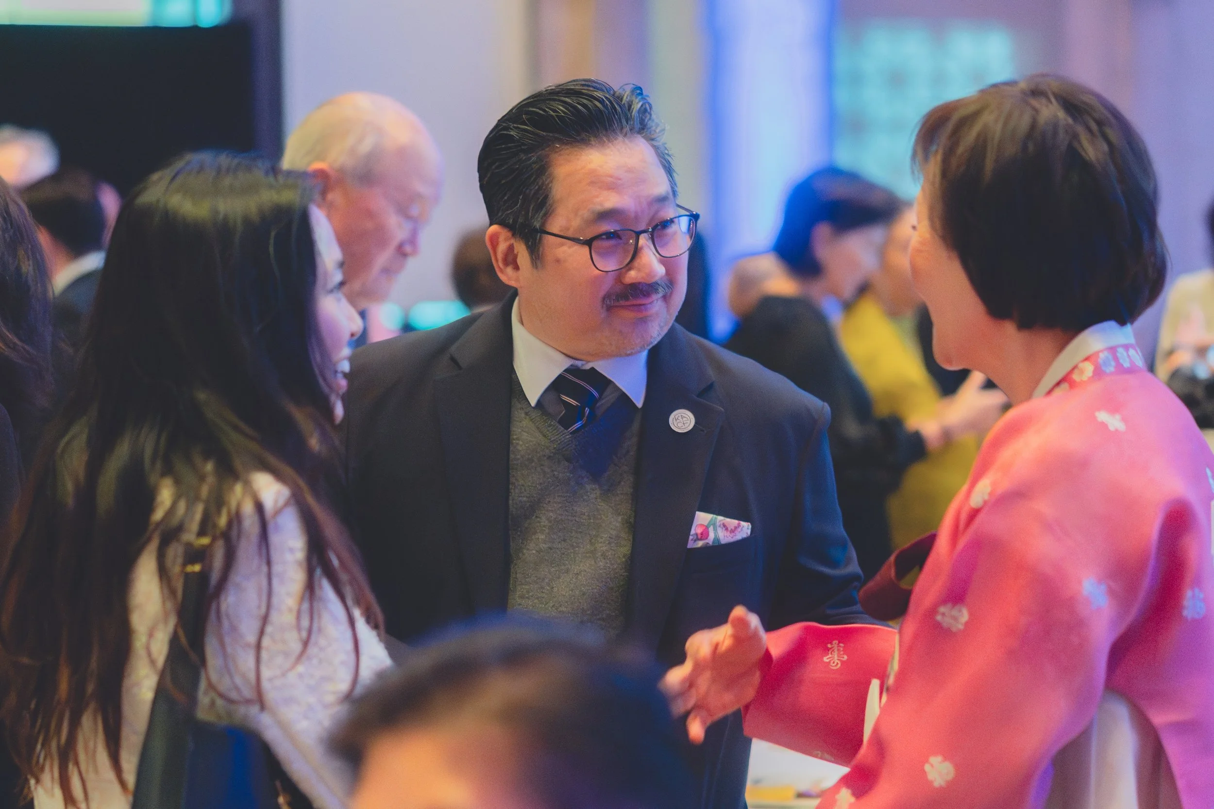 People at a formal event, a man with glasses and a woman in a pink kimono shaking hands.