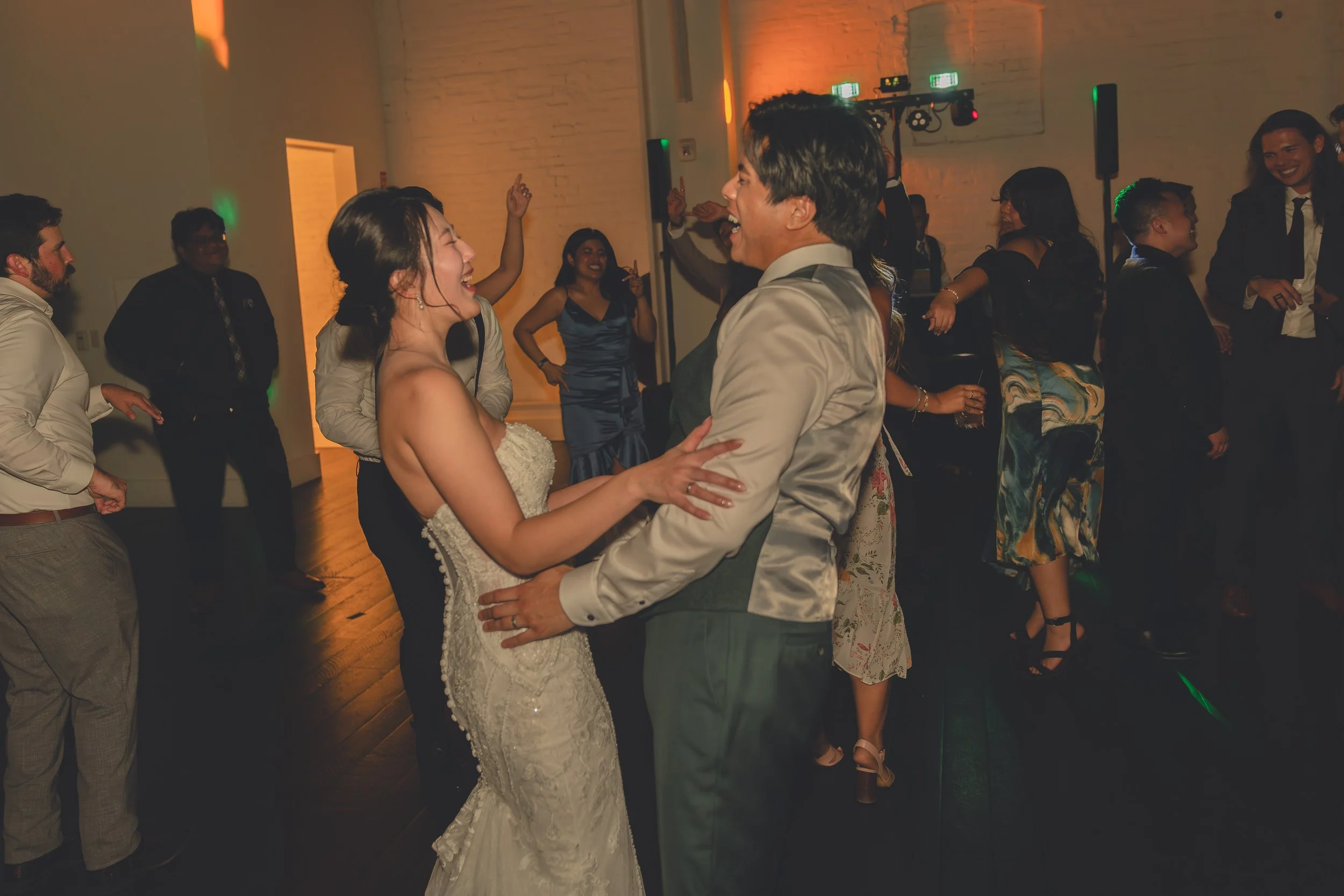 Wedding reception dance floor with a bride and groom dancing and smiling surrounded by guests in semi-formal attire in a dimly lit room.