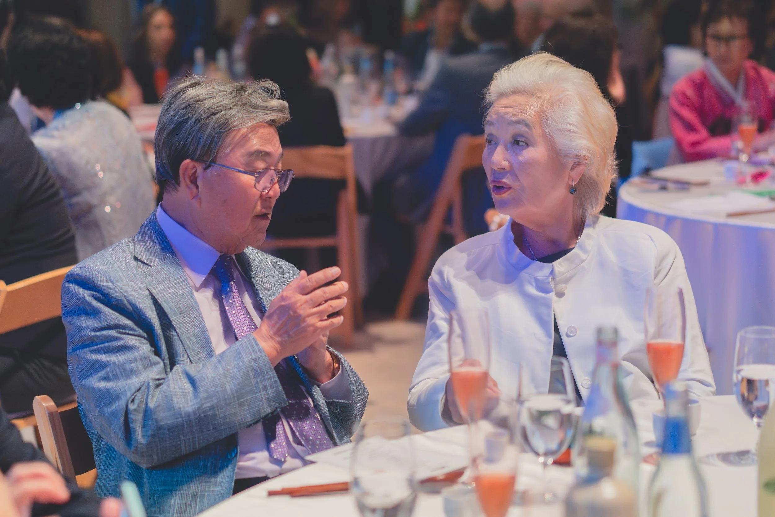 Two elderly women sitting at a banquet table engaged in conversation, with other guests in the background.