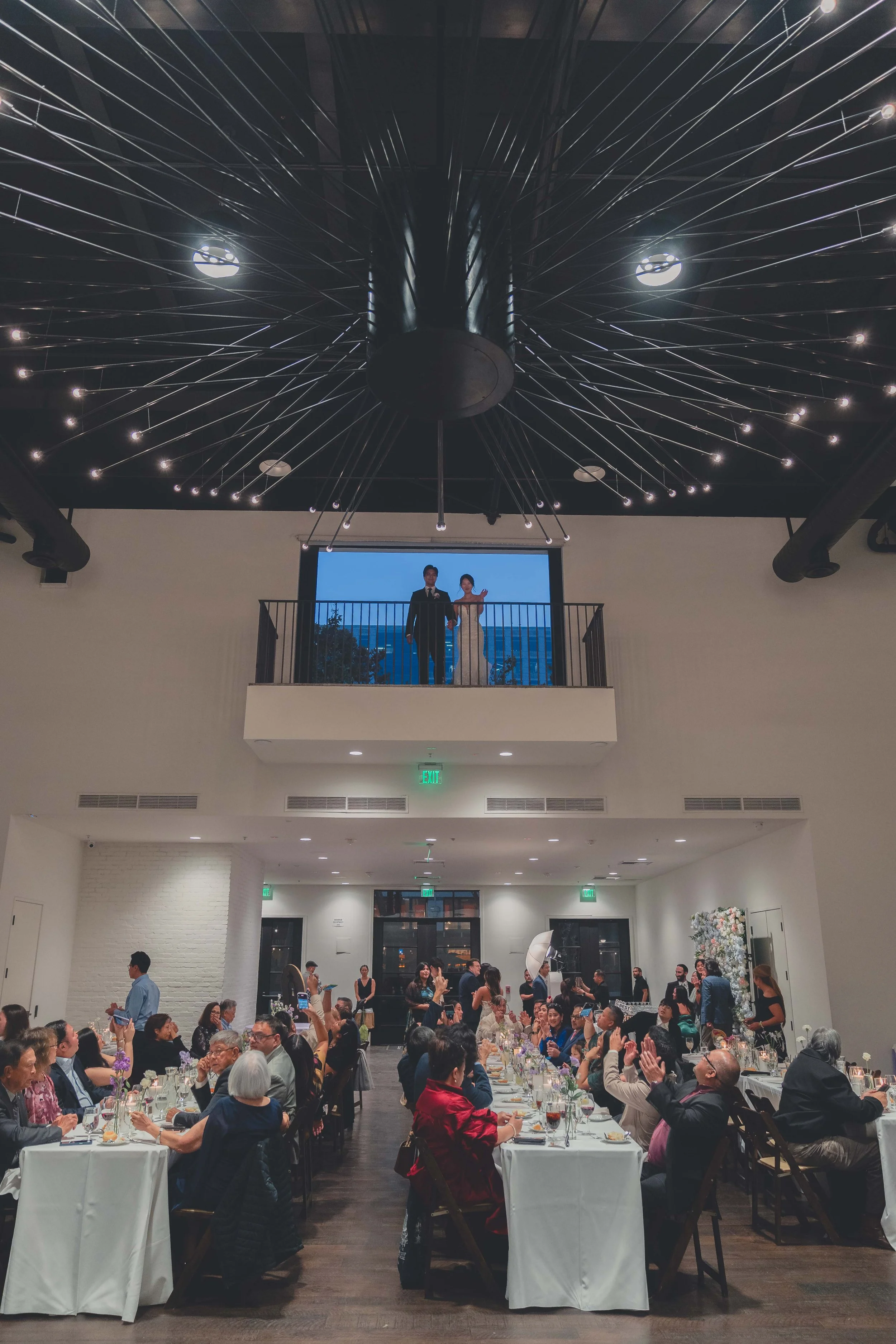 Wedding reception inside a hall with guests seated at tables, a couple standing on a balcony, and a decorative chandelier overhead.
