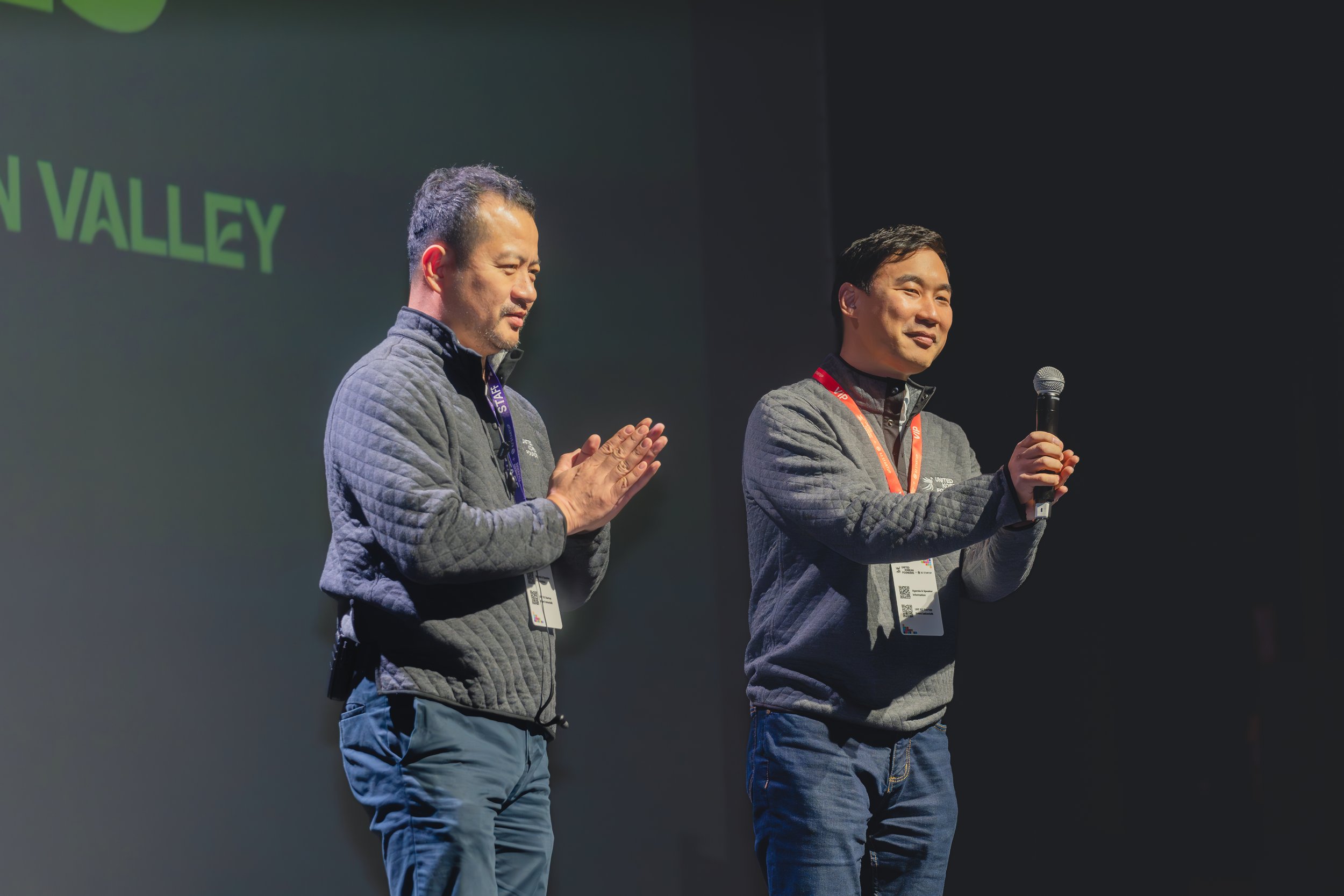 Two men standing on stage, one holding a microphone, wearing casual jackets, with conference name tags, against a dark background.