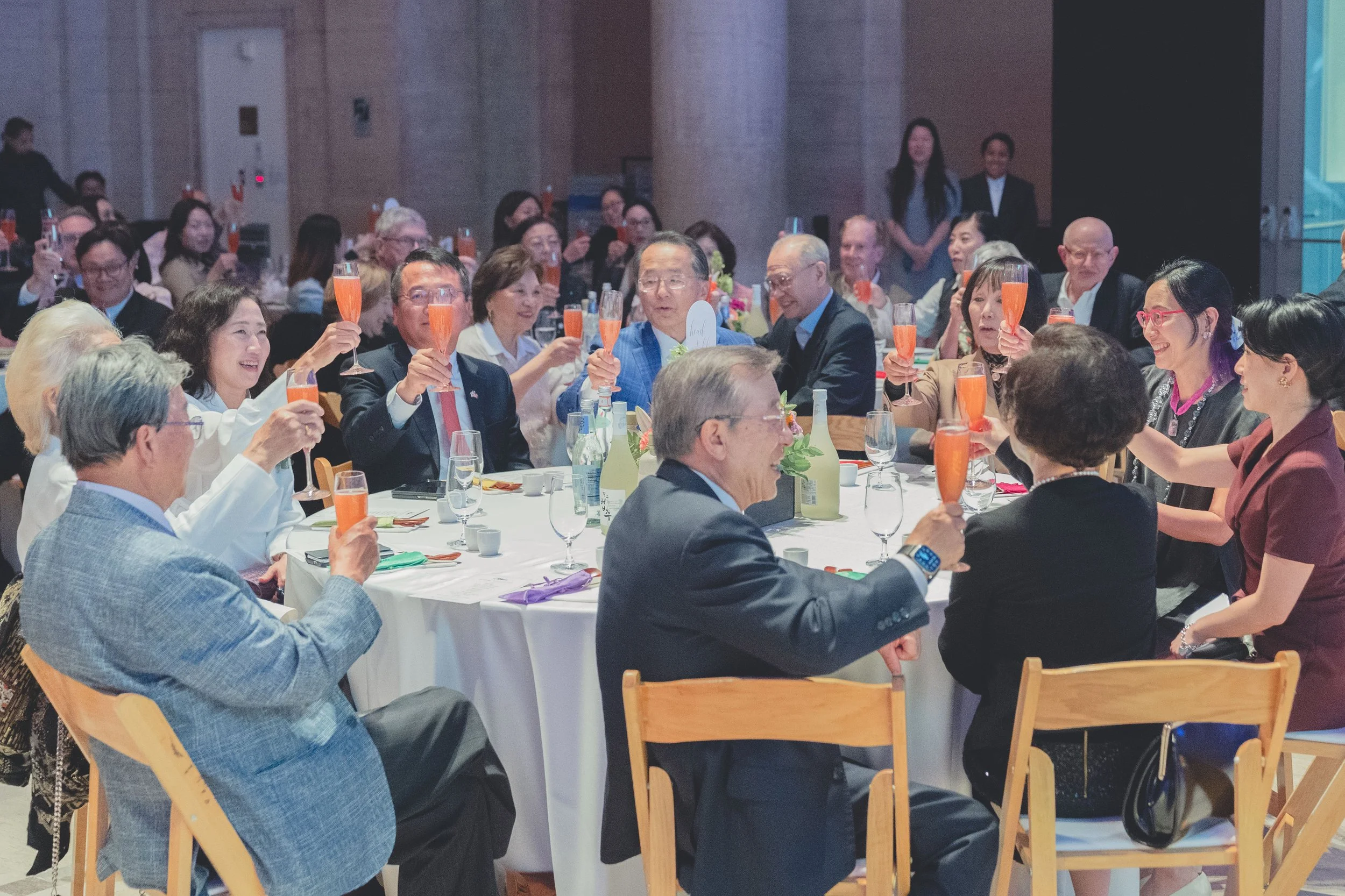 Gathering of diverse people seated around a large round table, celebrating with pink or orange sparkling drinks, in a formal event setting, with glasses, bottles, and table decorations.