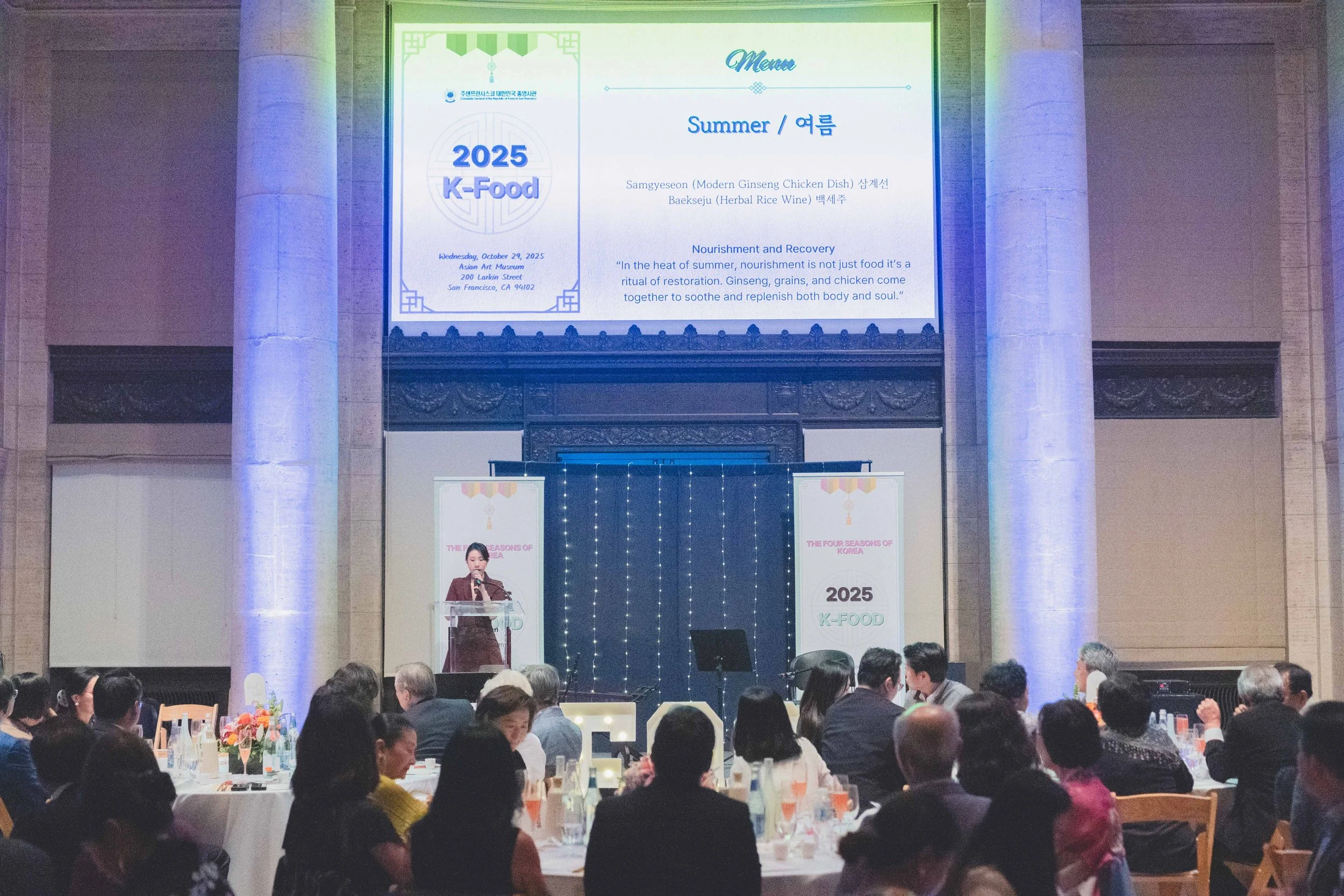 A large banquet hall with people seated at round tables, a woman speaking at a podium on stage, and a large screen displaying event details in Korean and English for the 2025 K-Food event, with decorative lighting and banners.