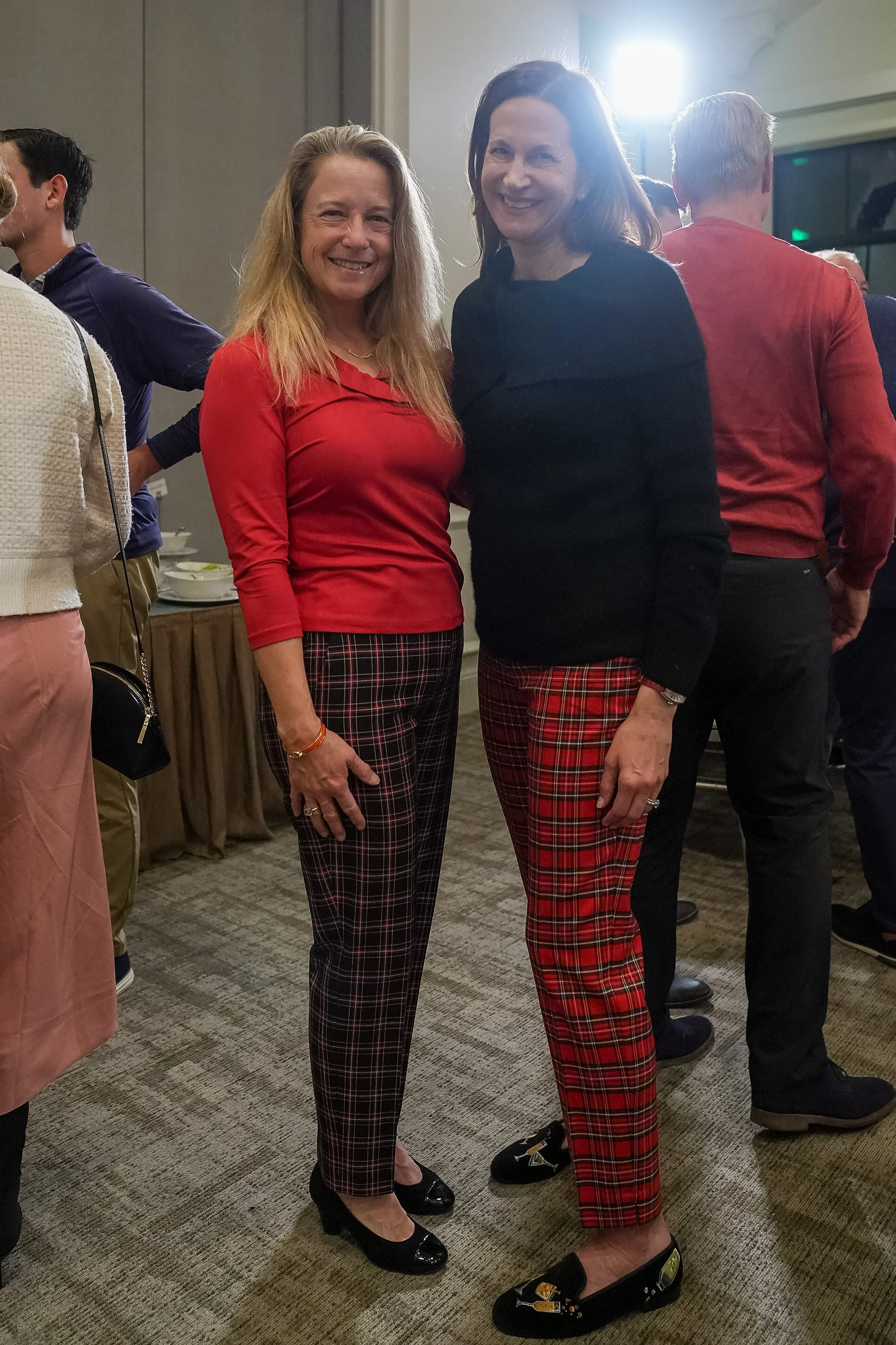 Two women smiling at a gathering, wearing plaid pants; one in a red top and the other in a black top.